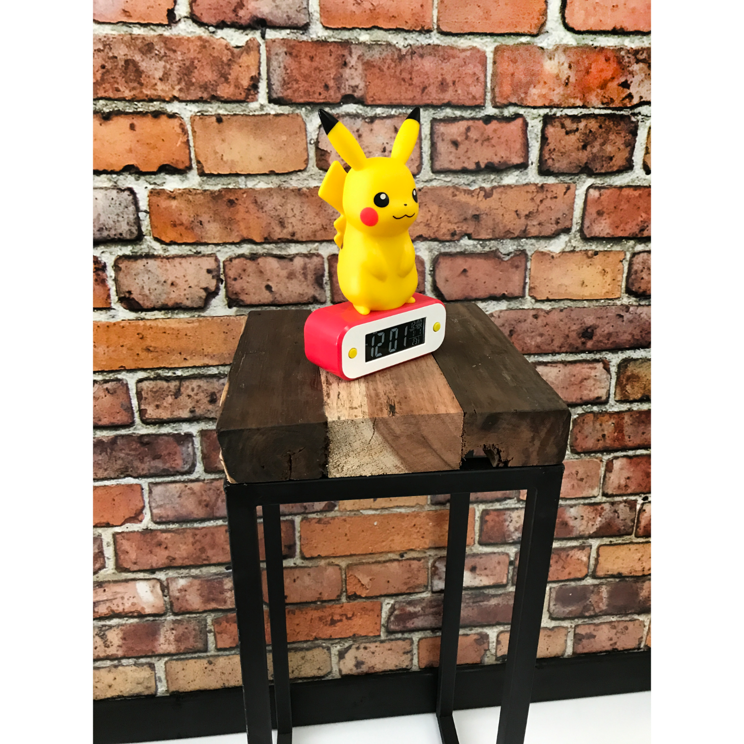 Figura di Pikachu su sveglia rossa e bianca, su supporto in legno e metallo, muro di mattoni.