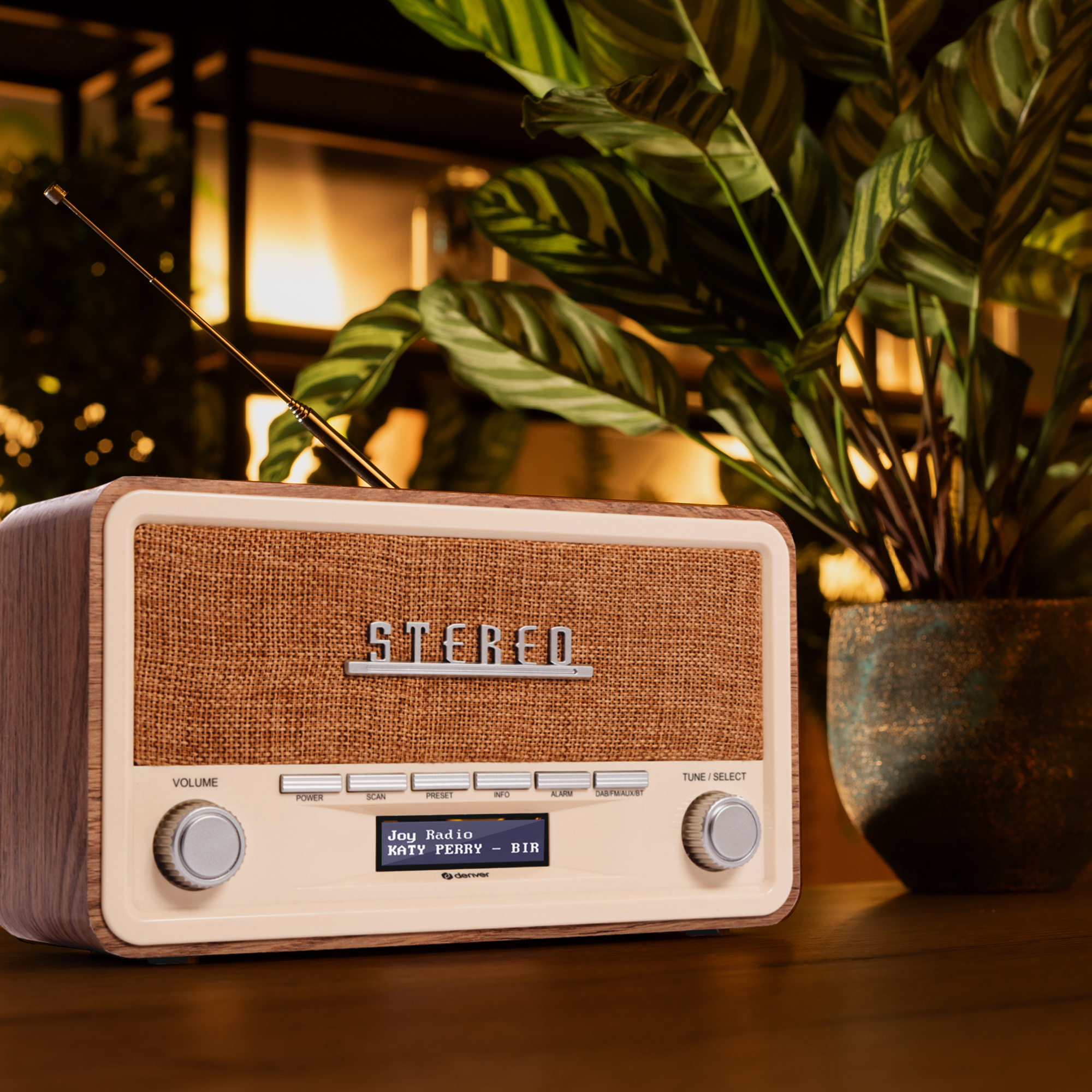 A retro radio with a brown speaker, wood trim, and a small display, placed near a plant.