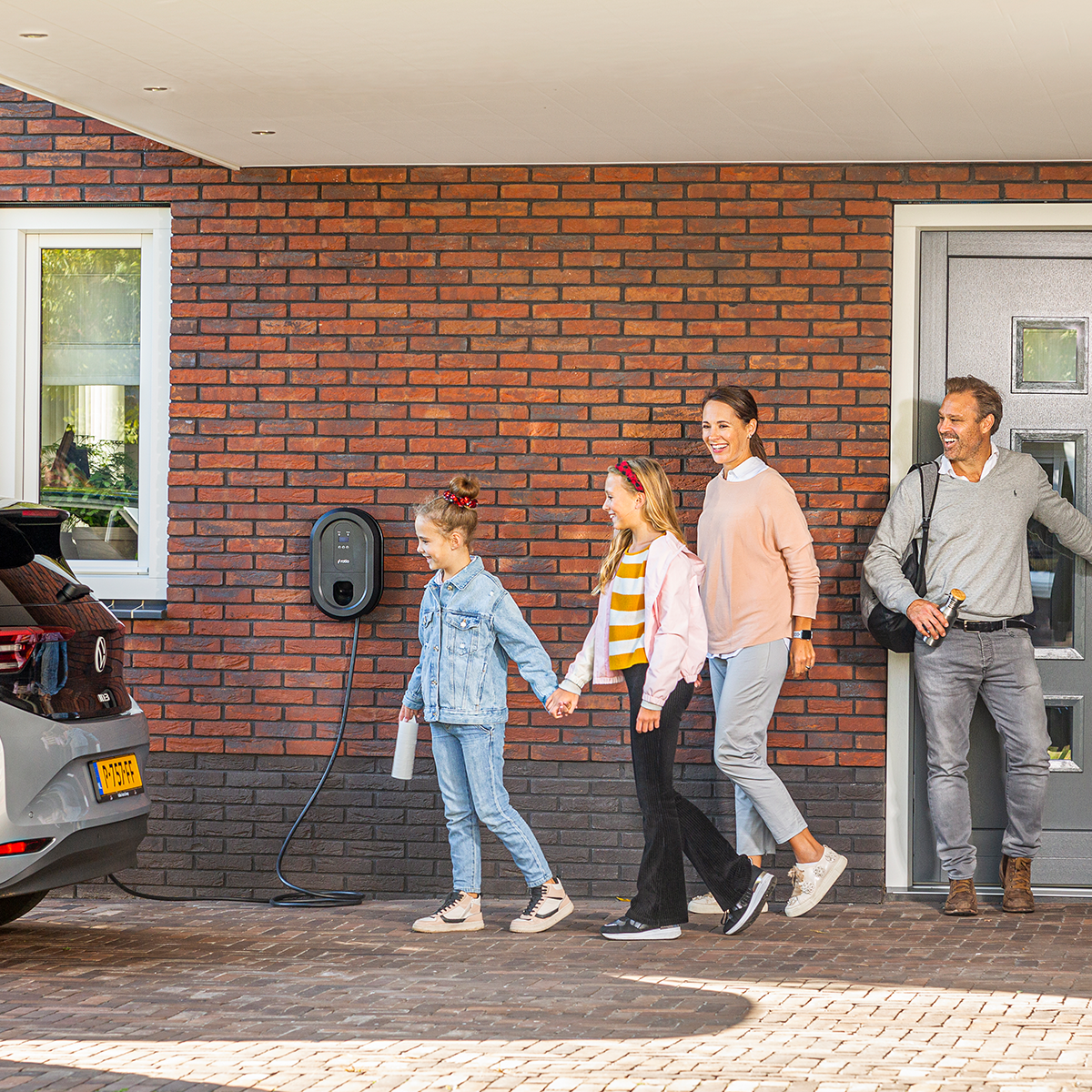 Family walking towards a house with an electric car charging. Wall charger on the wall.