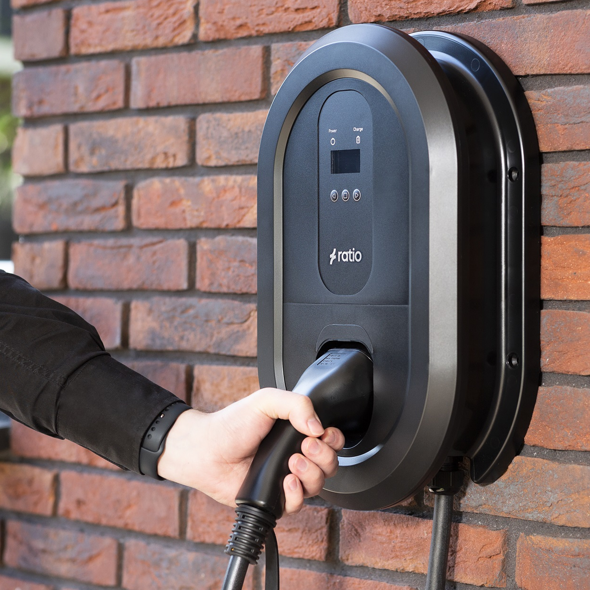 Black Ratio EV charger mounted on a brick wall. Hand holding charging connector.