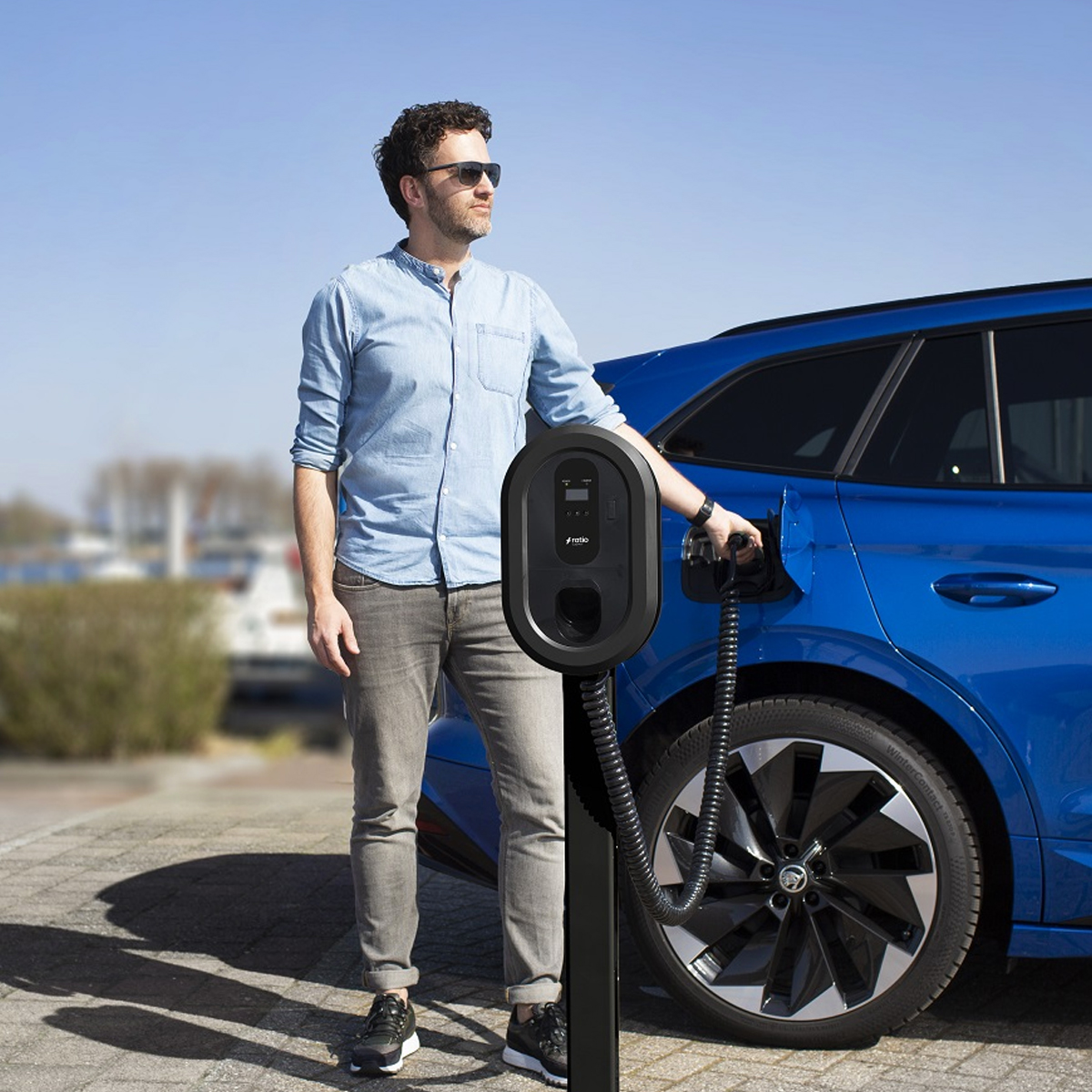 Man in sunglasses charging a blue car at an electric charging station. Sunny day, blue sky.