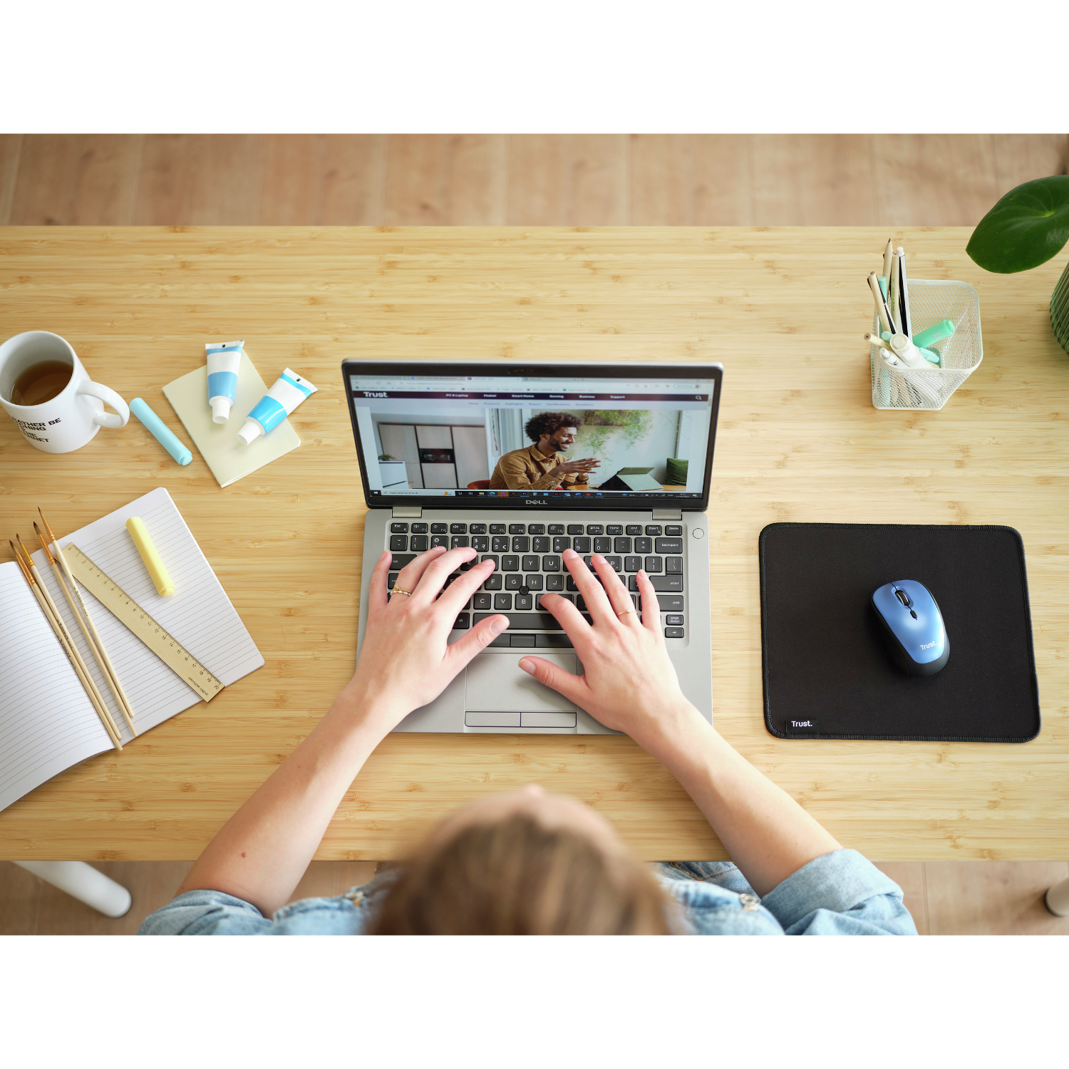 Persona che lavora su un laptop a una scrivania. C'è un mouse, penne, una tazza e tubetti di vernice.