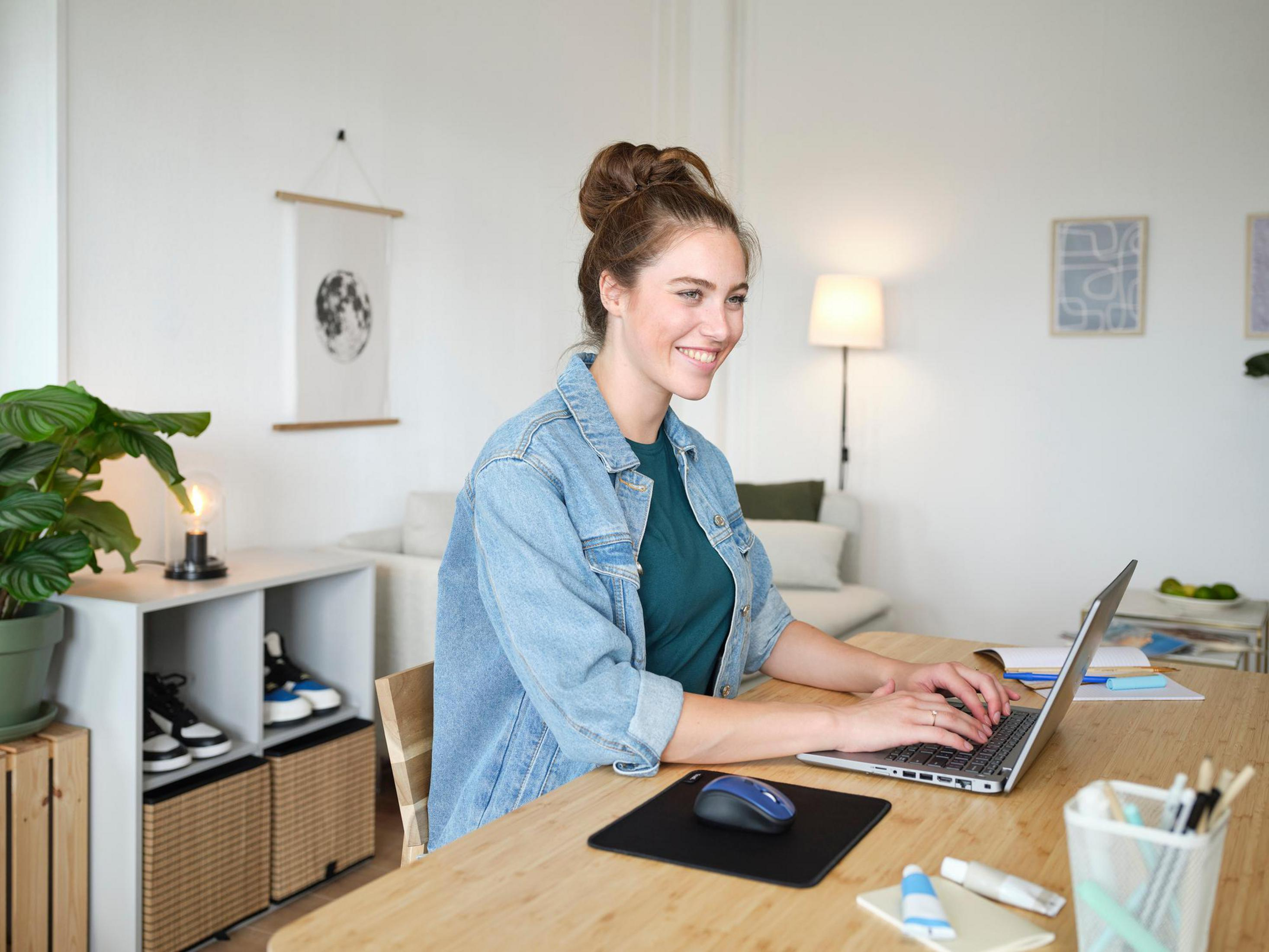 Donna con giacca di jeans che lavora su un laptop a una scrivania in legno. Stanza con piante e opere d'arte.