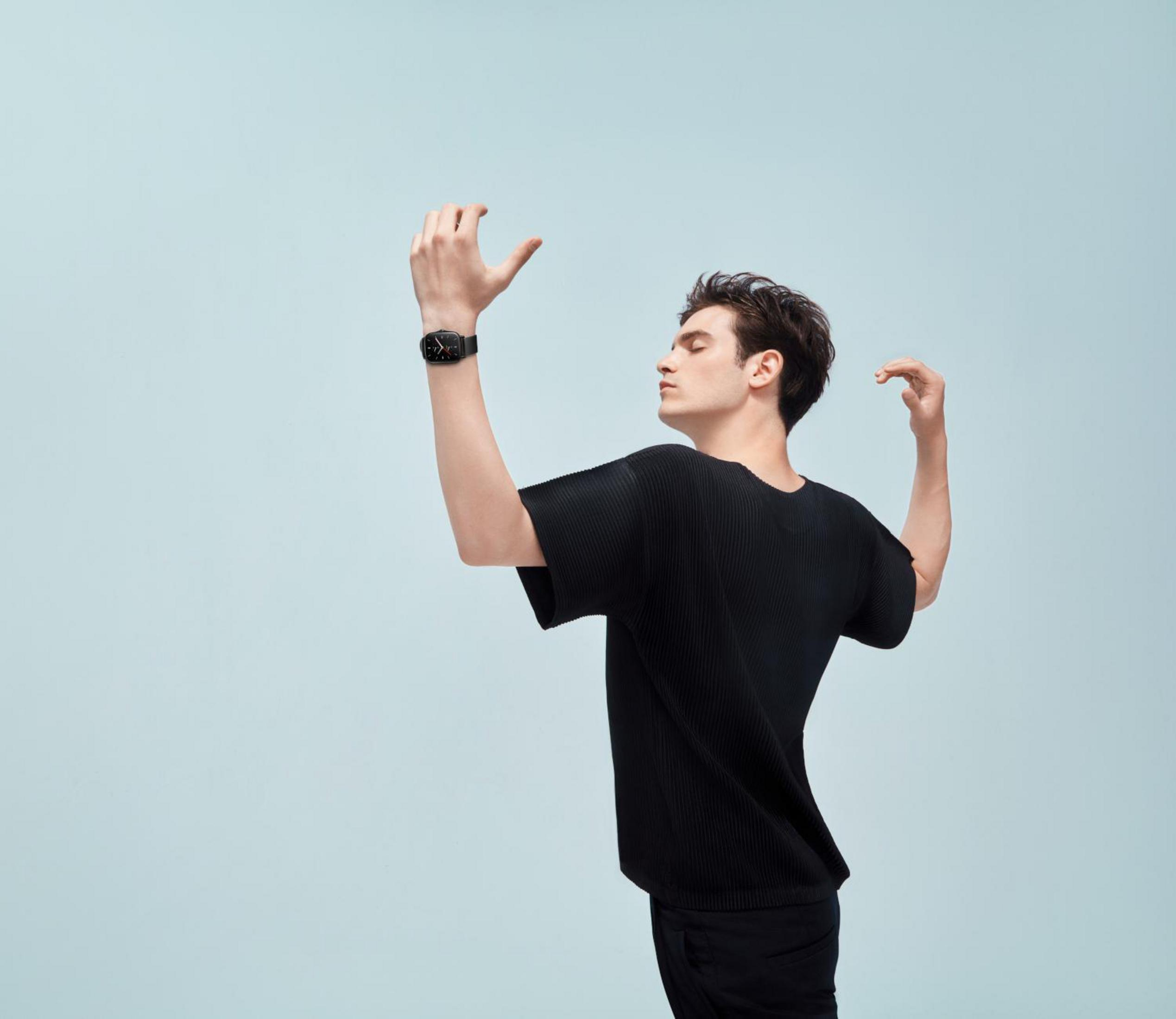 Mężczyzna w czarnej koszuli i zegarku, z podniesionymi ramionami, stojący na jasnoniebieskim tle.