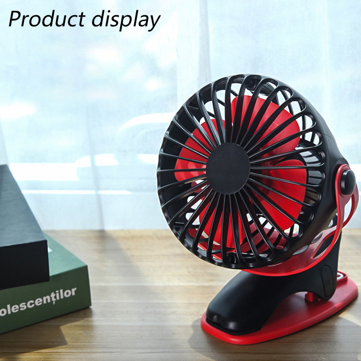 Ein rot-schwarzer Ventilator auf einem Schreibtisch mit einem grünen Buch, Fenster im Hintergrund.
