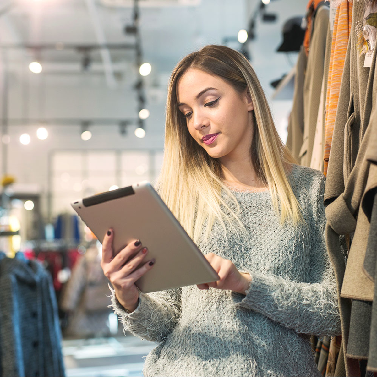 Kobieta z blond włosami patrząca na tablet w sklepie odzieżowym. W tle stojaki na ubrania.