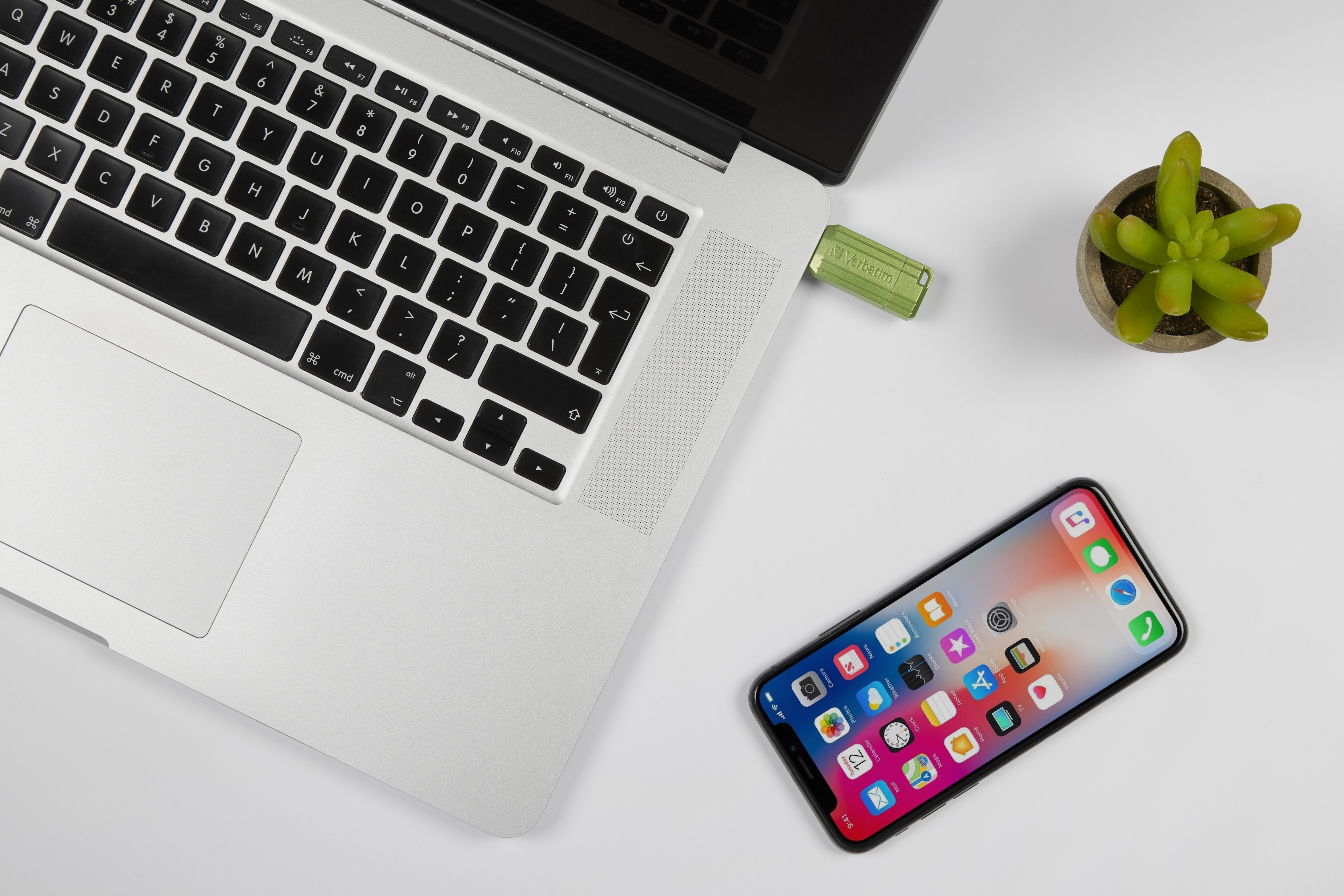 Un laptop, una chiavetta USB, un telefono e una piccola pianta sono su una superficie bianca.