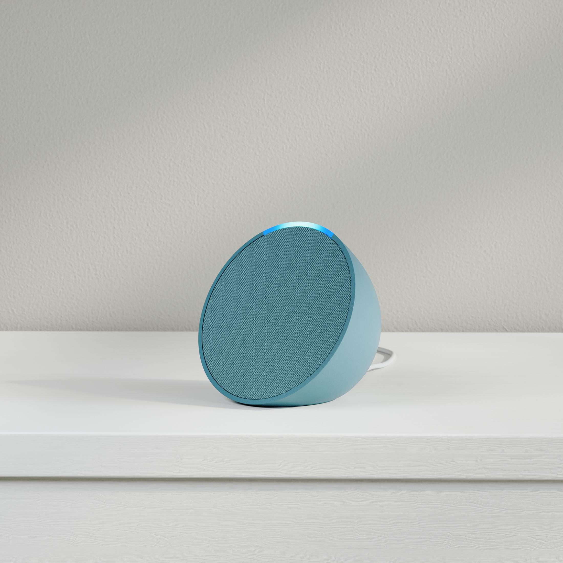 Głośnik w kolorze turkusowym na białej powierzchni. Na górze ma niebieskie światło.