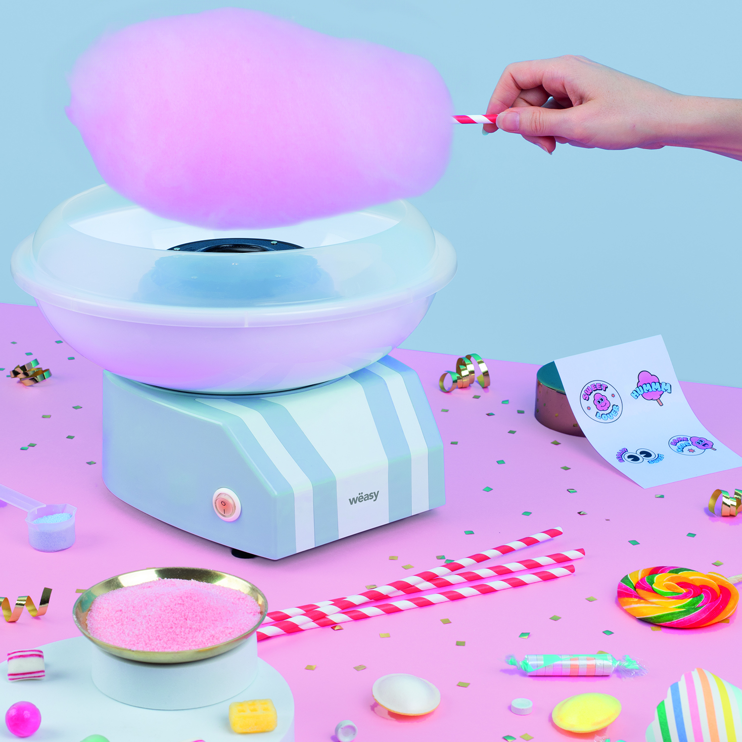 Zuckerwattemaschine mit rosa Zuckerwatte, umgeben von Süßigkeiten auf rosa Oberfläche.