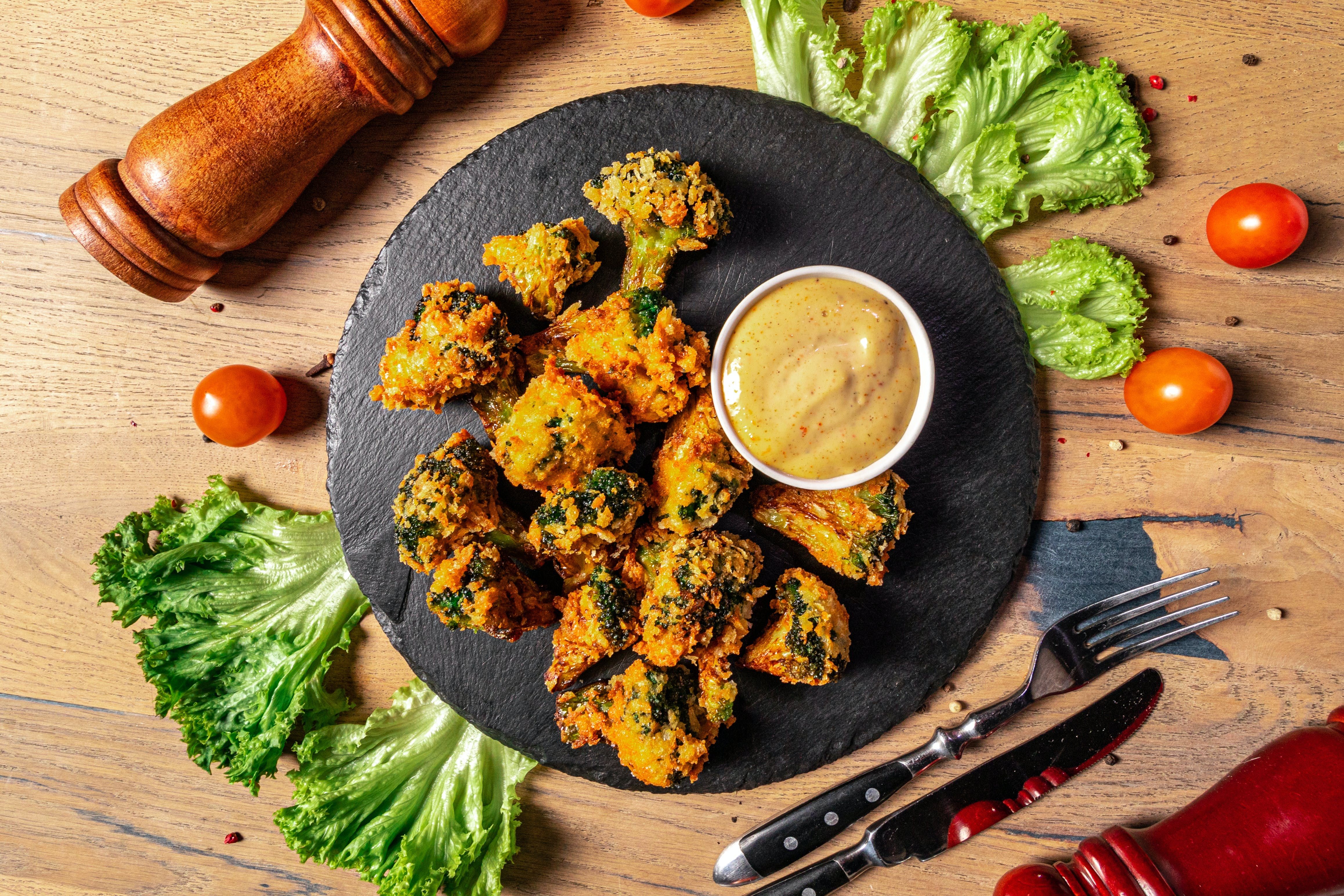 Gebakken broccoli op een zwart bord met een dipsaus. Tomaten, sla en een pepermolen staan ​​in de buurt.