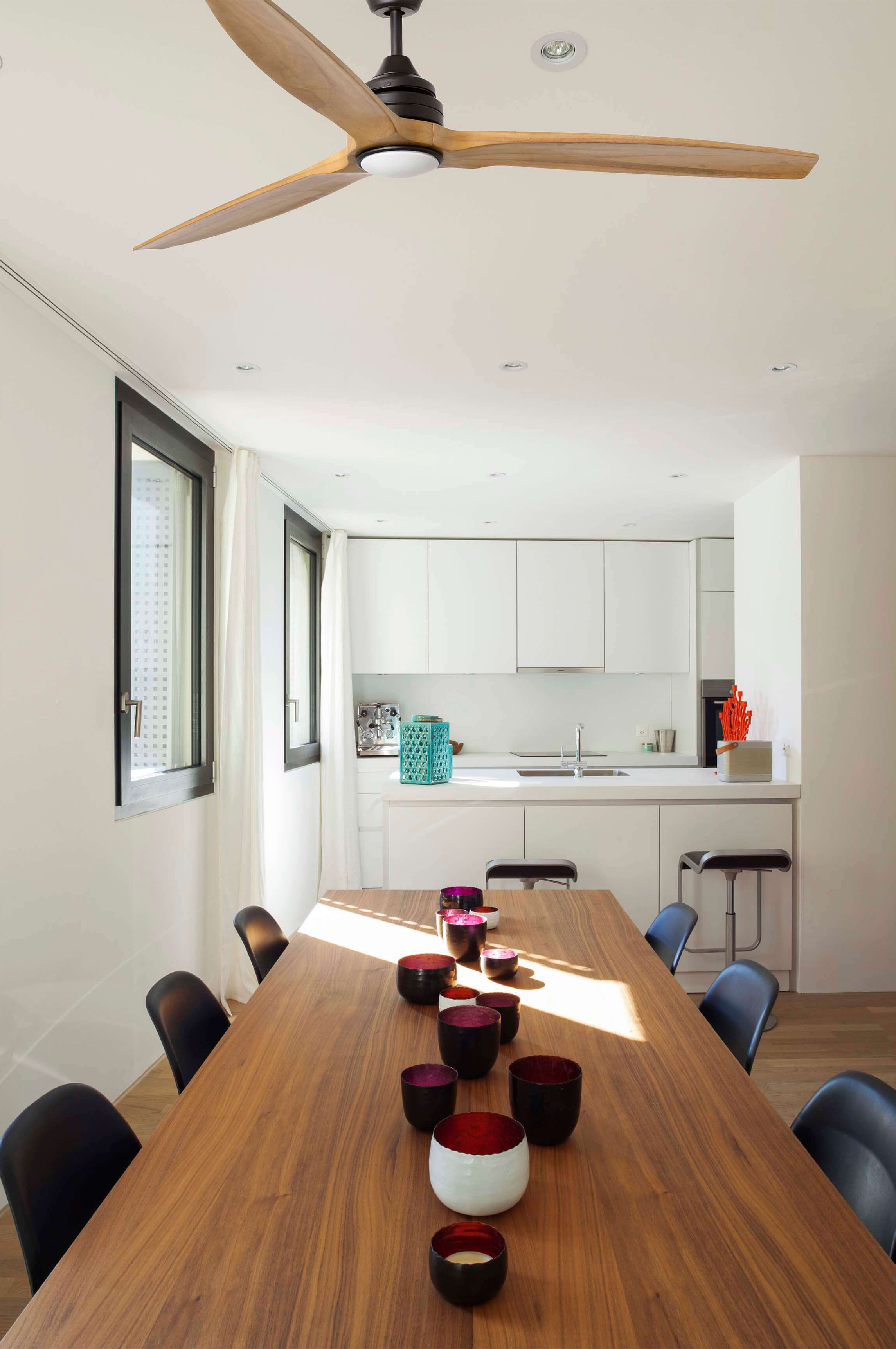 Sala da pranzo con tavolo in legno, sedie nere, cucina bianca e ventilatore a soffitto. Oggetti decorativi.