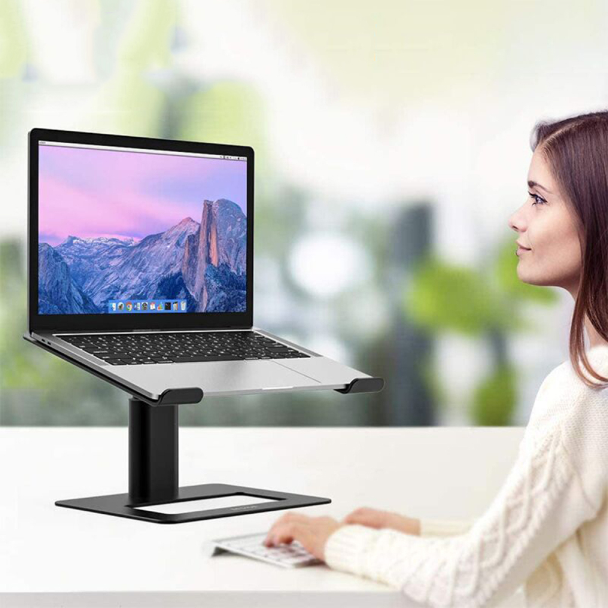 Eine Frau sitzt an einem Schreibtisch mit einem Laptop auf einem schwarzen Ständer und schaut auf den Bildschirm.
