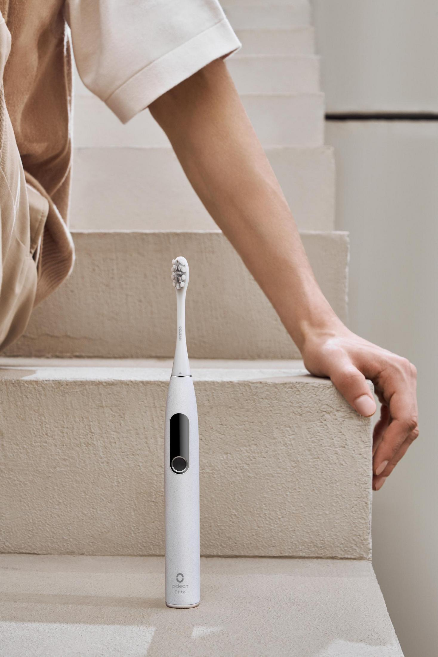 Ramię osoby spoczywa na schodach, trzymając białą elektryczną szczoteczkę do zębów. Czarny przycisk.