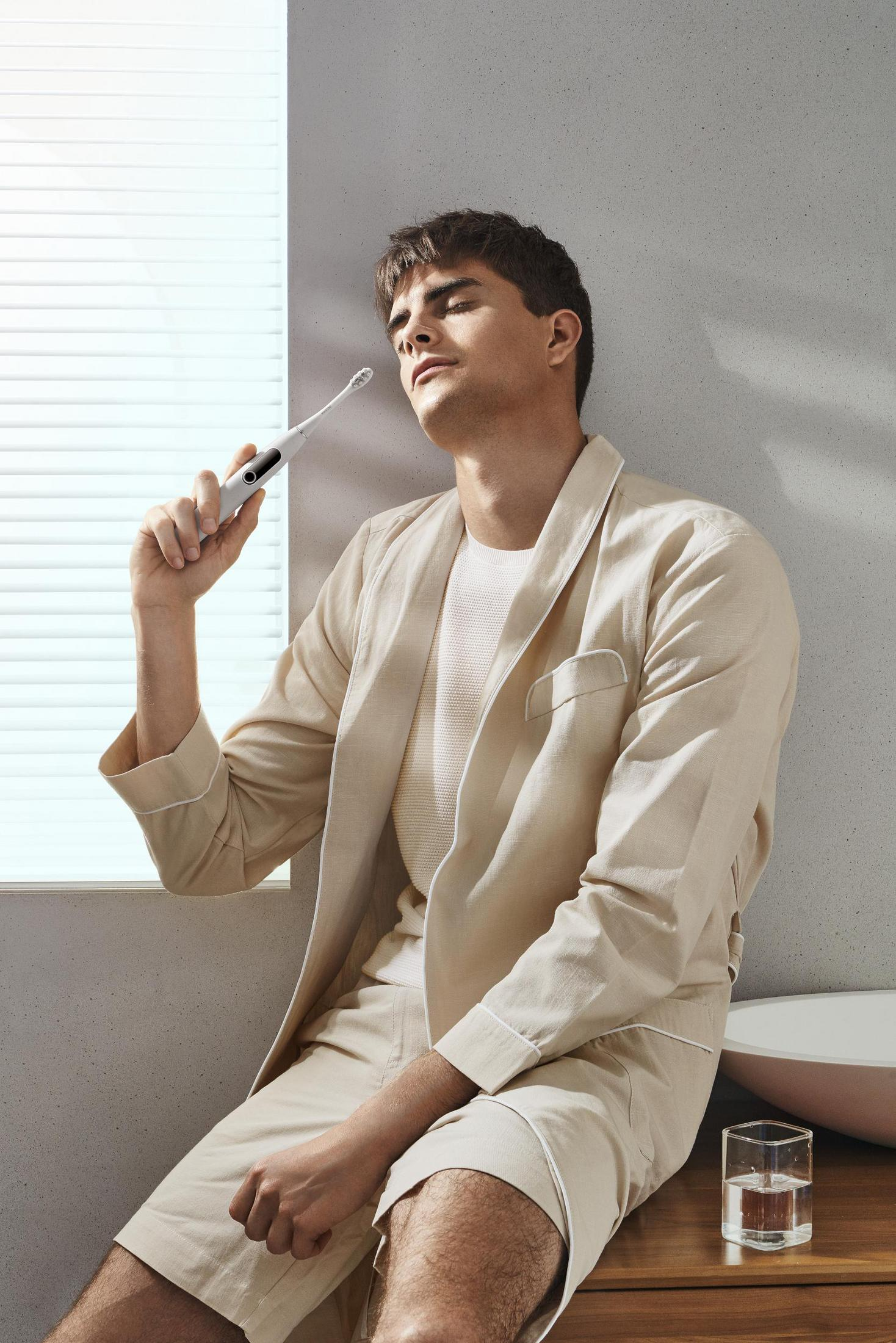 Mężczyzna w beżowym szlafroku trzymający białą elektryczną szczoteczkę do zębów, patrzy w górę.