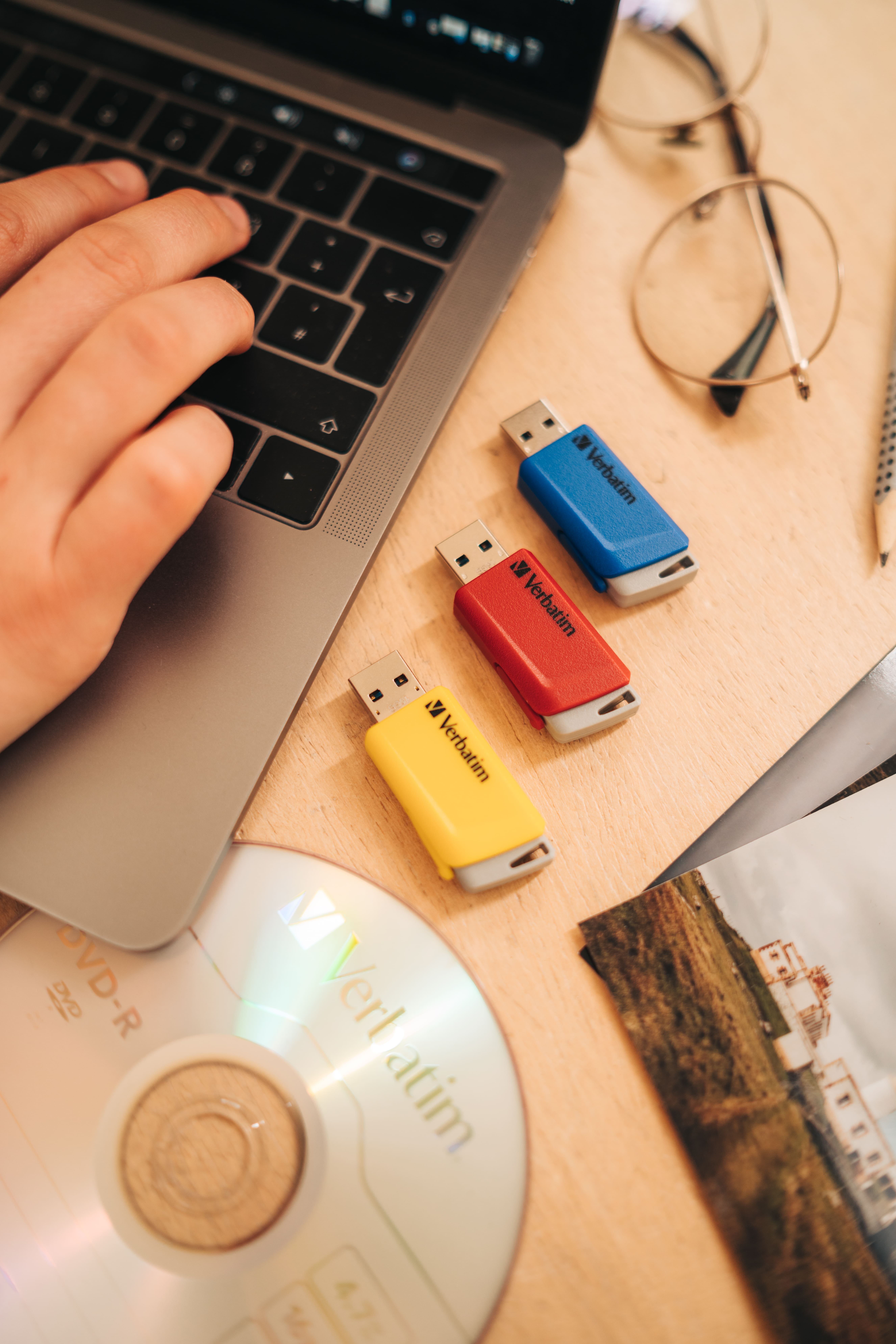 Konfiguracja biurka z laptopem, trzema pendrive'ami, płytą CD, okularami i długopisem. Na drewnianej powierzchni.