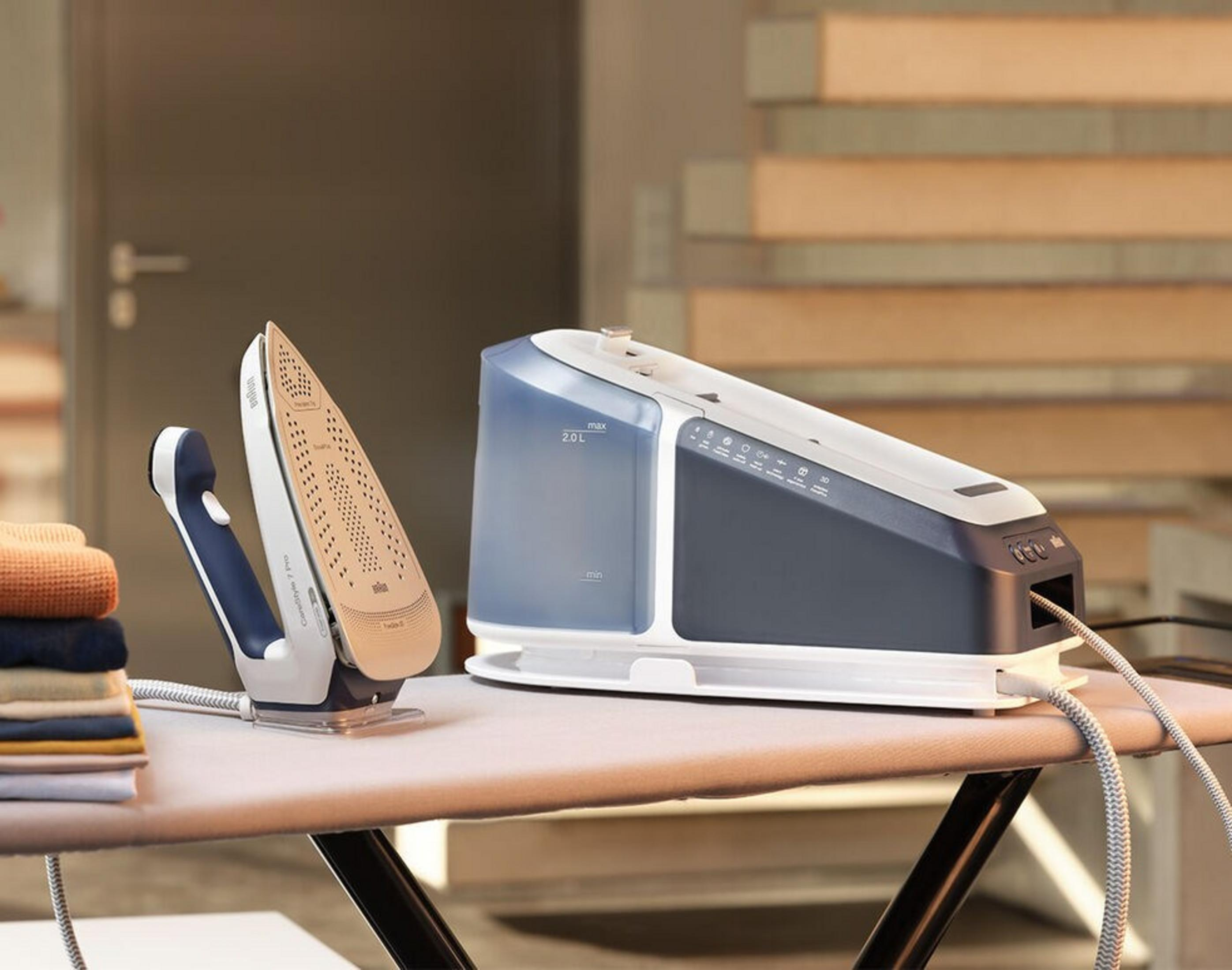 Ironing station on ironing board. Iron, water tank, and stack of clothes are visible.