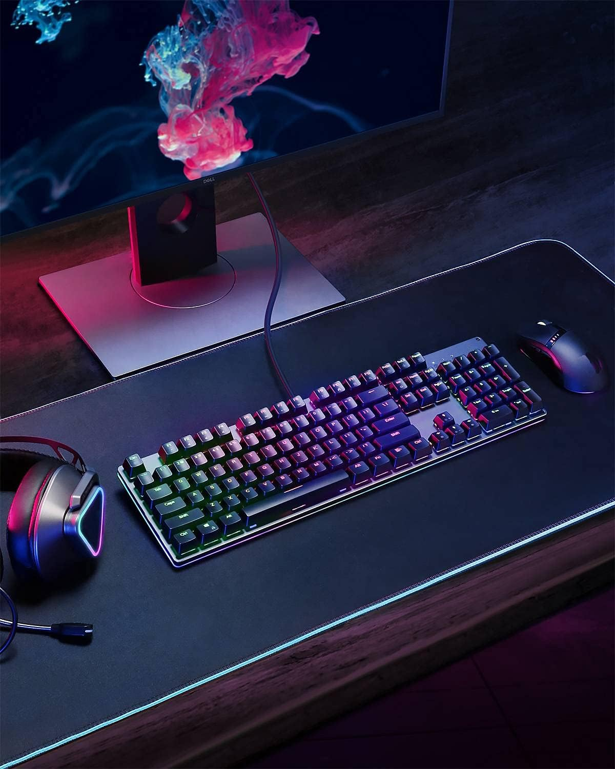 Configuración de juego con teclado, ratón, auriculares y monitor. El teclado tiene luces RGB. El fondo está borroso.