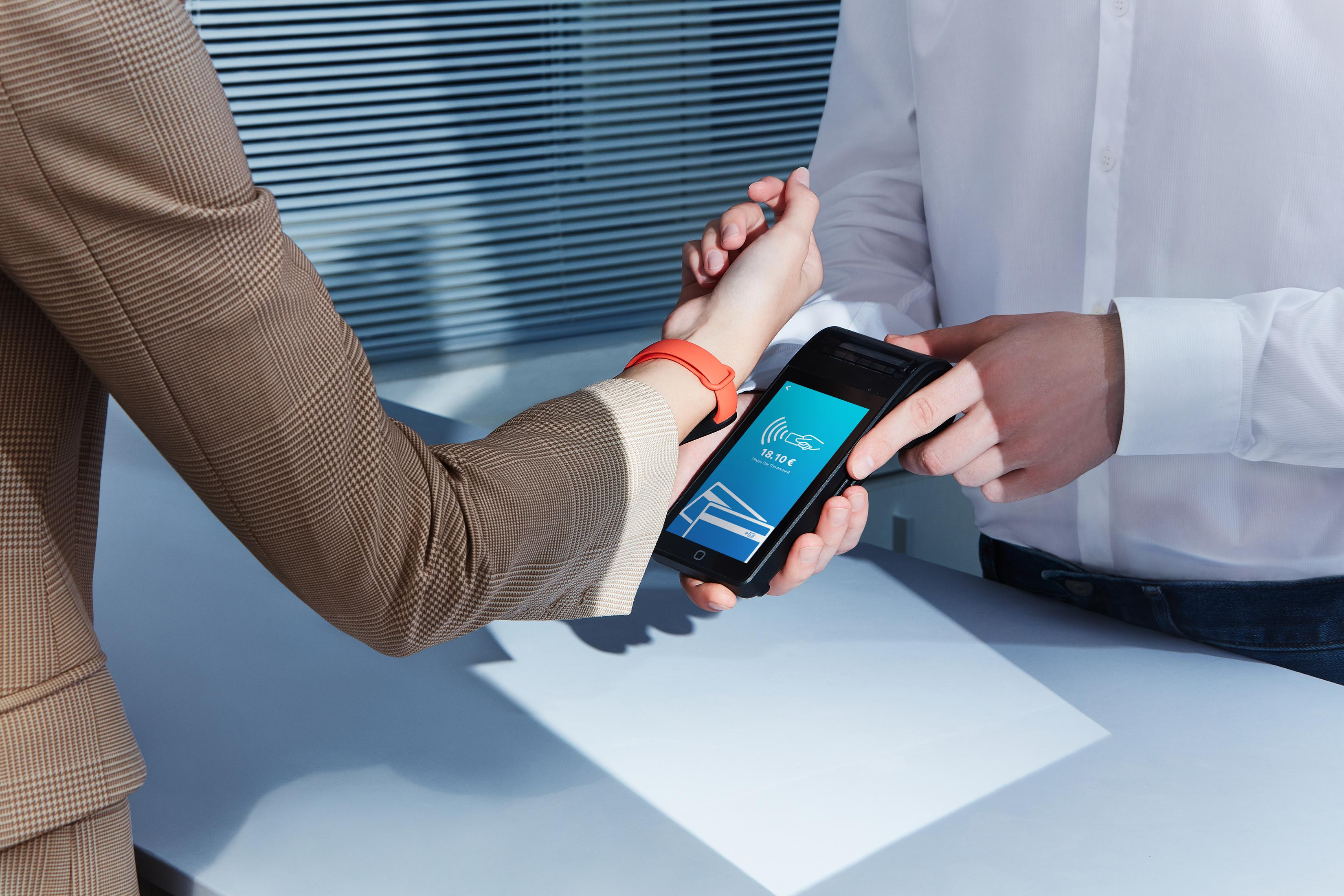 Eine Person zahlt mit einem Telefon in der Nähe des Handgelenks einer Person mit orangefarbenem Armband, drinnen.