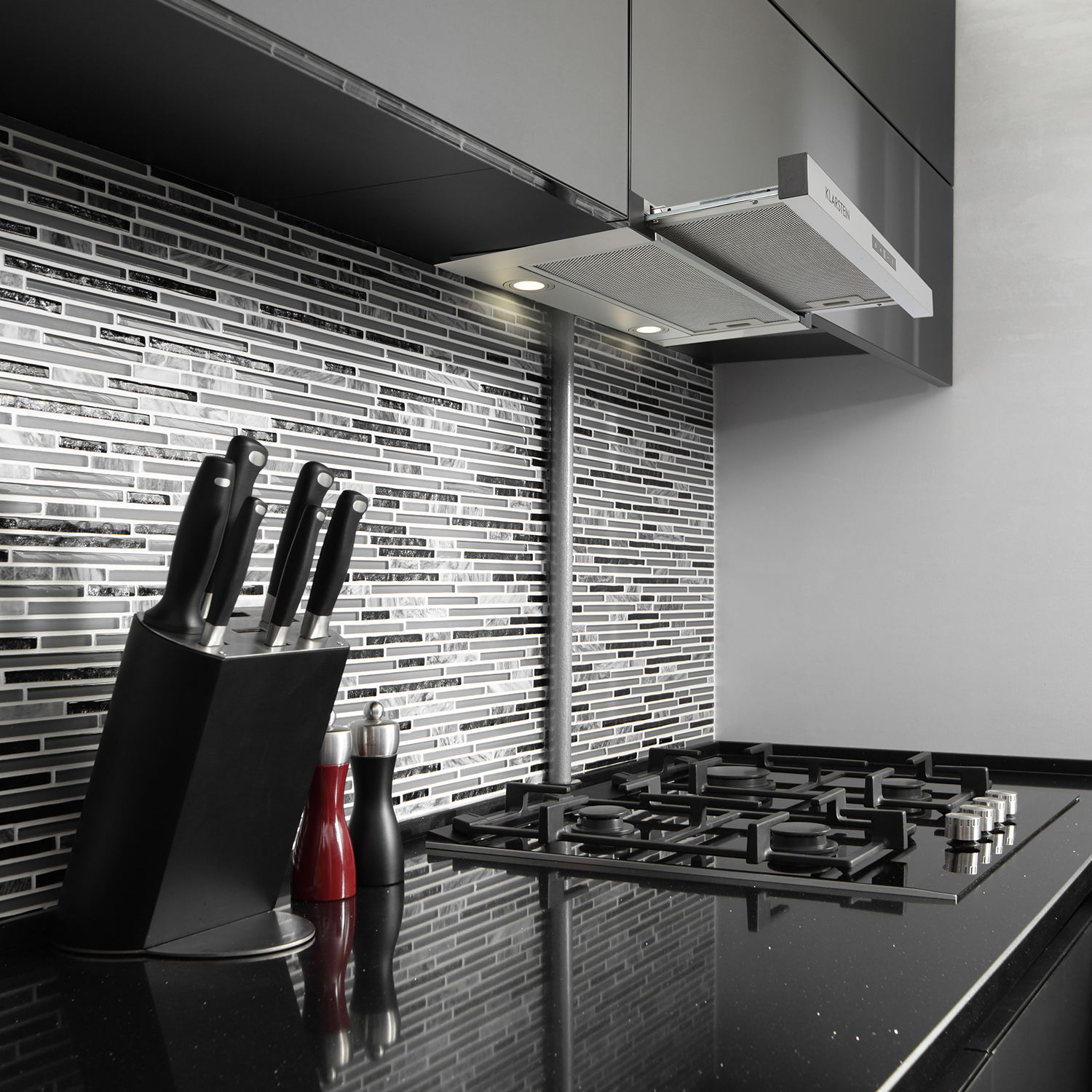 Una escena de cocina con encimera negra, una estufa y una campana extractora con luces. Azulejos blancos y negros.