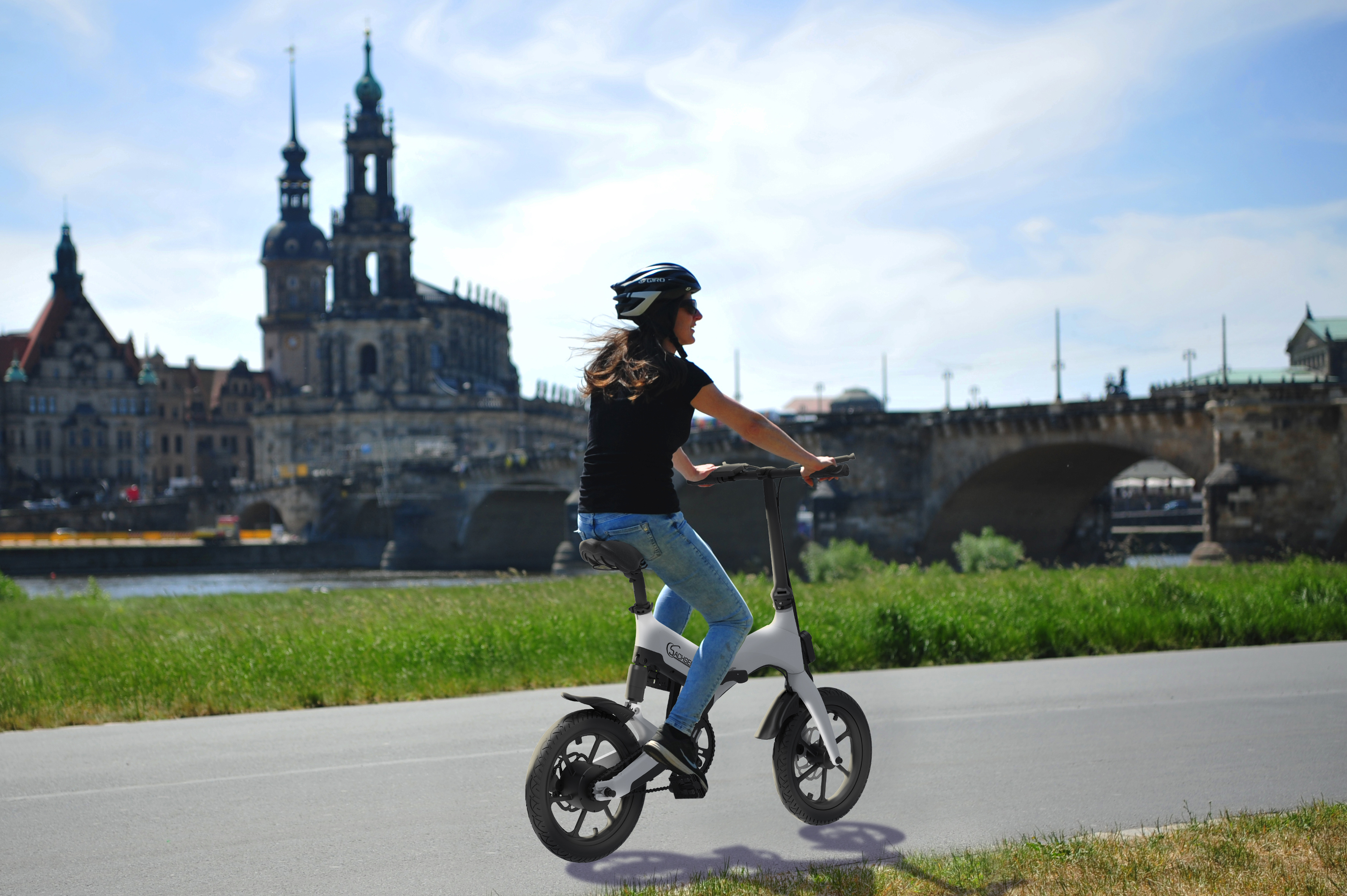 Frau fährt mit einem weißen Elektrofahrrad auf einer Straße. Im Hintergrund eine Stadt mit einer Brücke.