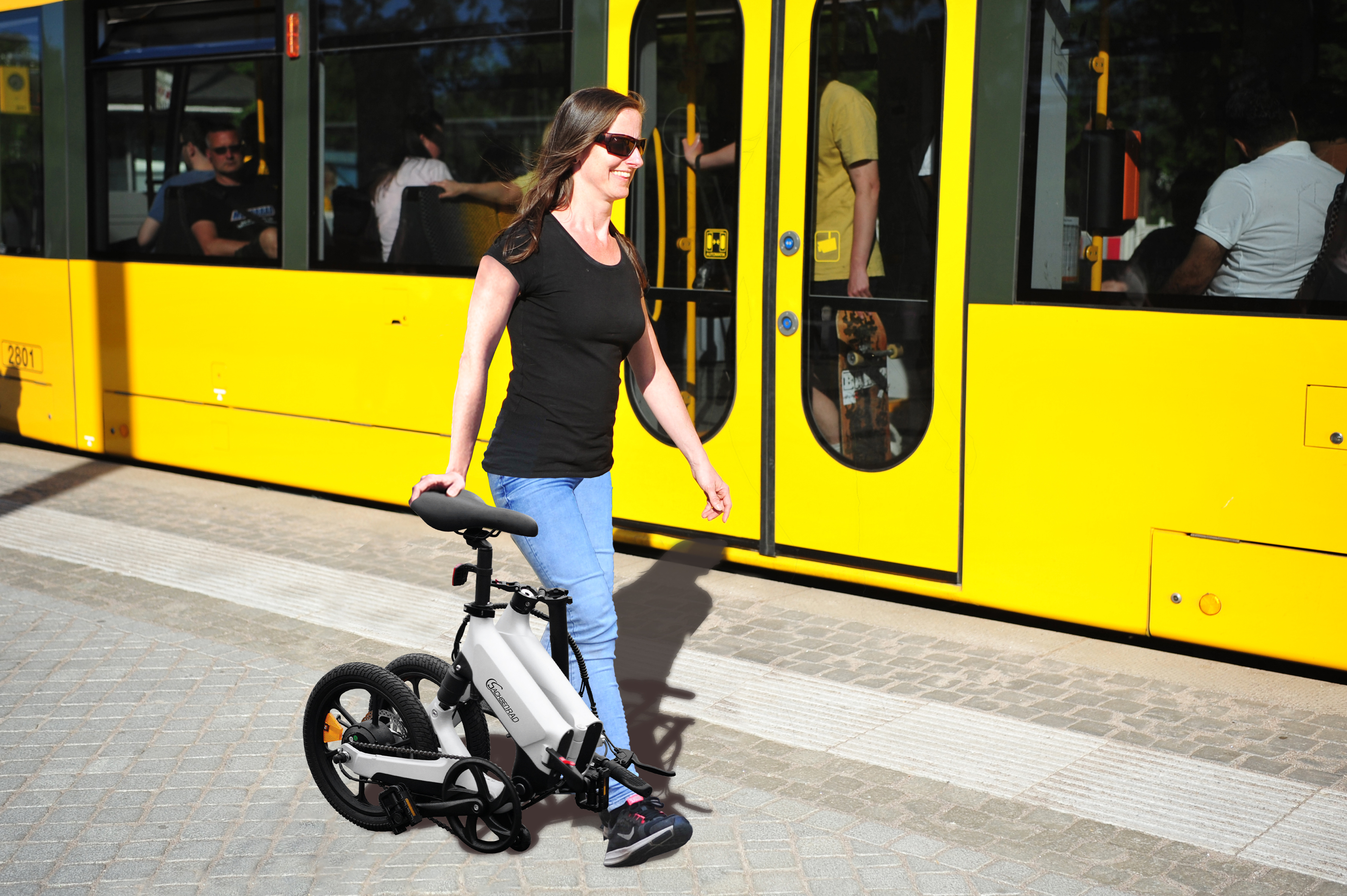 Frau mit Fahrrad, geht an einem gelben Bus vorbei. Sonniger Tag.