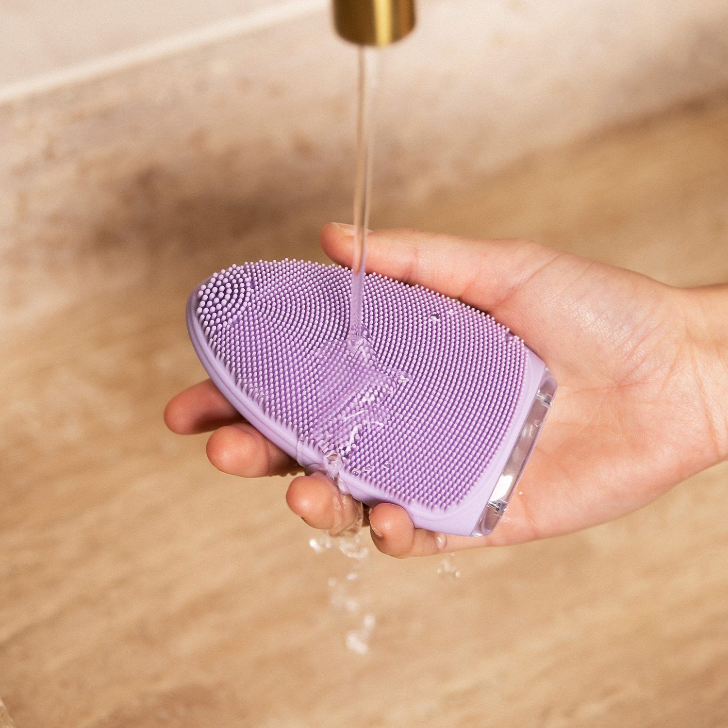 Eine Hand hält eine lilafarbene Gesichtsreinigungsbürste unter einem Wasserstrahl.