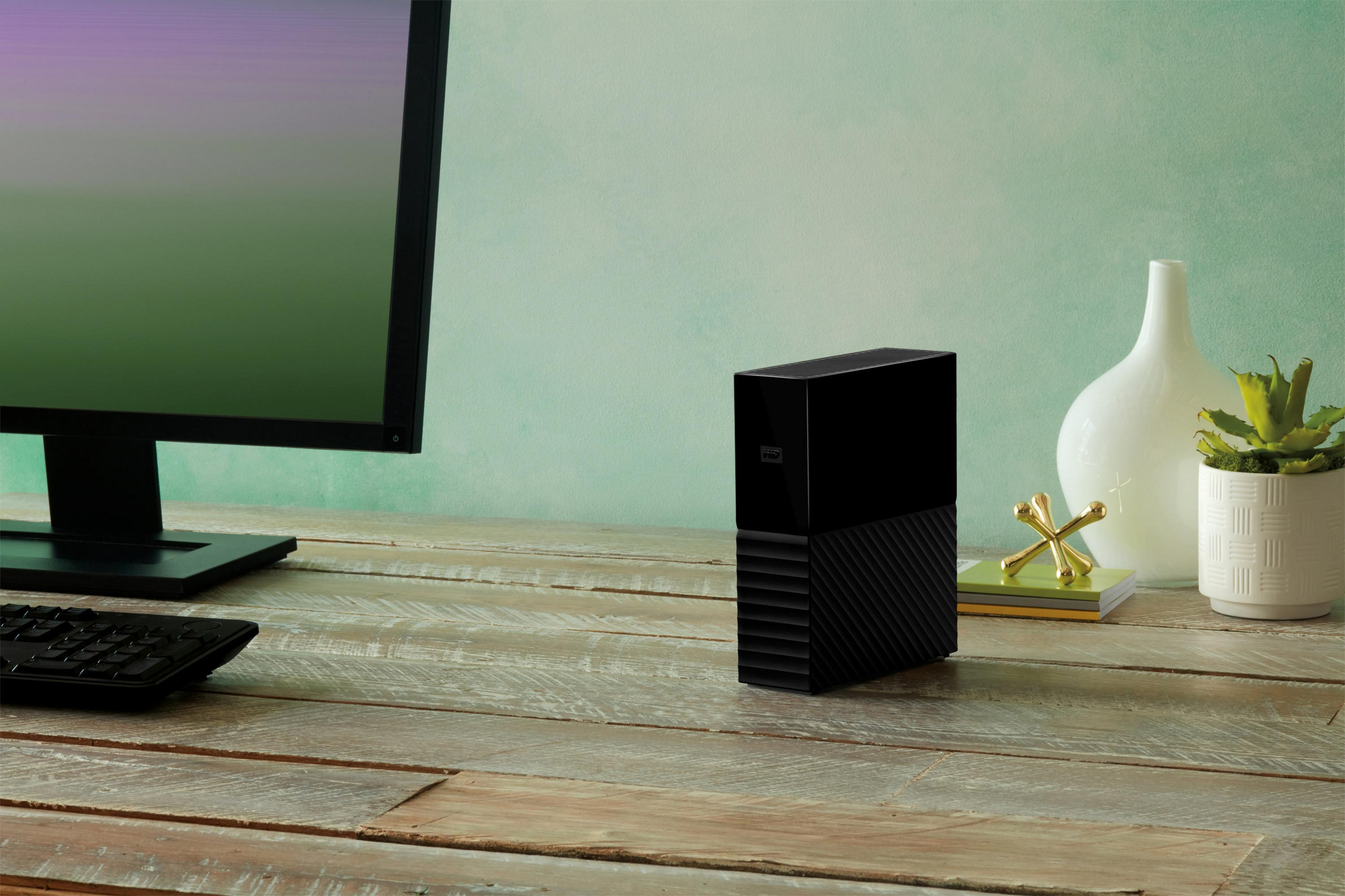 Czarny zewnętrzny dysk twardy stoi na drewnianym biurku obok monitora i dekoracji, z zieloną ścianą.