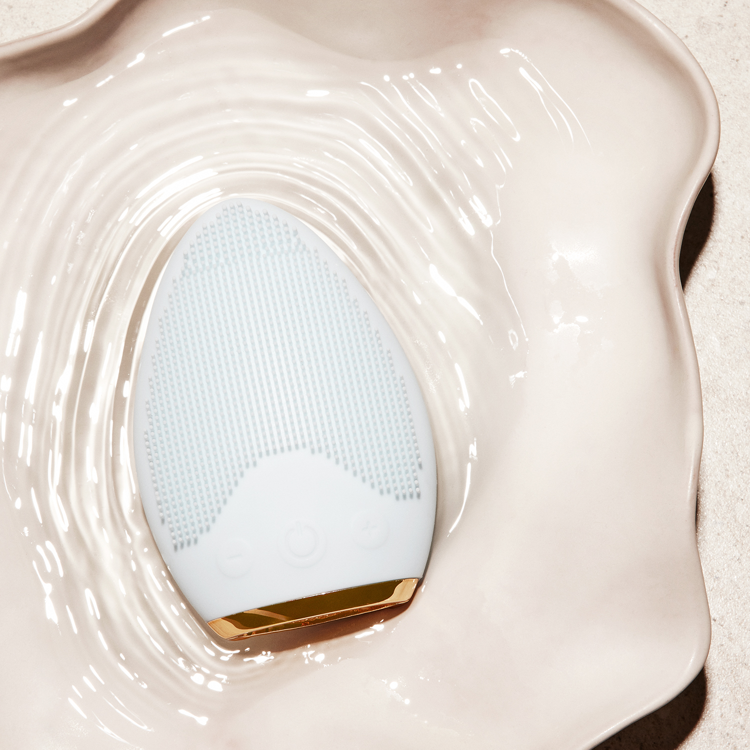 Weiß-goldene Gesichtsreinigungsbürste in einer weißen, muschelförmigen Schale mit Wasser. Auf weißem Grund.