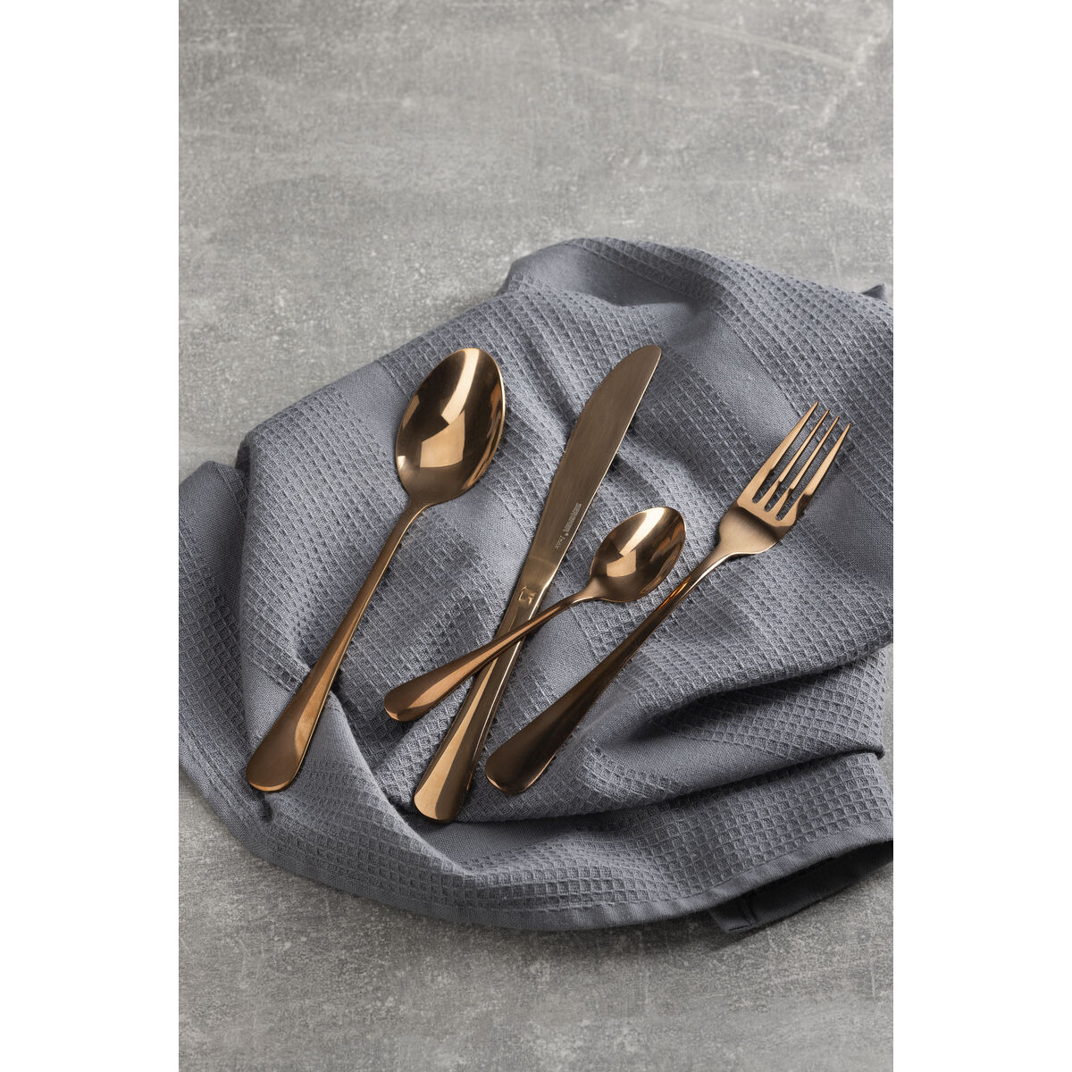 Cubertería color cobre sobre tela gris: cuchara, cuchara pequeña, cuchillo y tenedor.