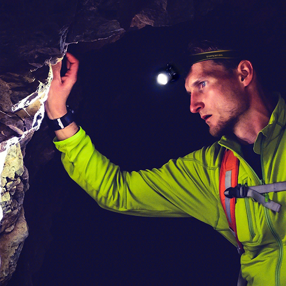 Mann in Höhle mit Stirnlampe, berührt Felswand, dunkler Hintergrund.