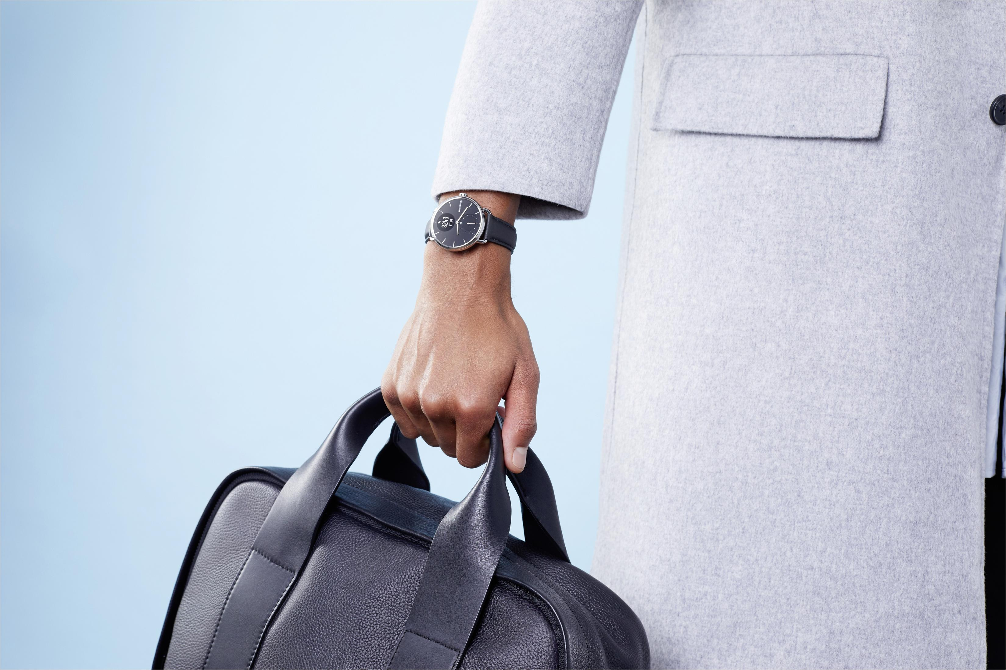 Person hält schwarze Tasche, trägt grauen Mantel und Armbanduhr, hellblauer Hintergrund.