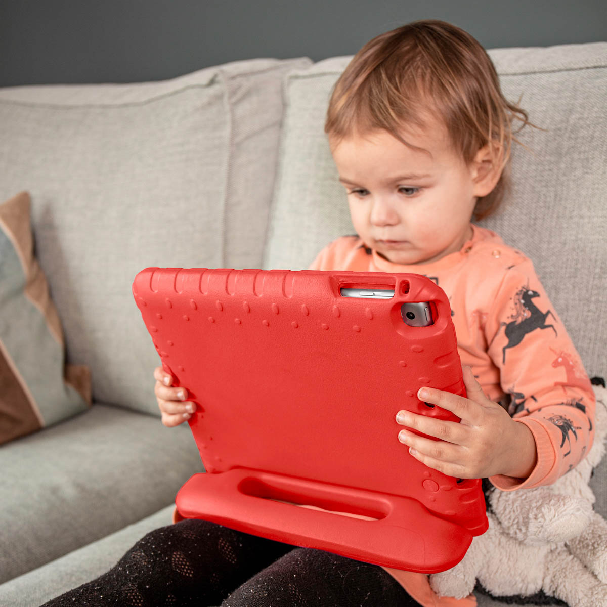 A young child sits on a couch, holding a red tablet case. The child is looking at the tablet.