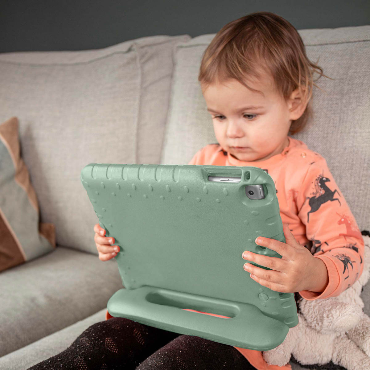 Een kind houdt een tablet in een groene hoes vast, zittend op een grijze bank.