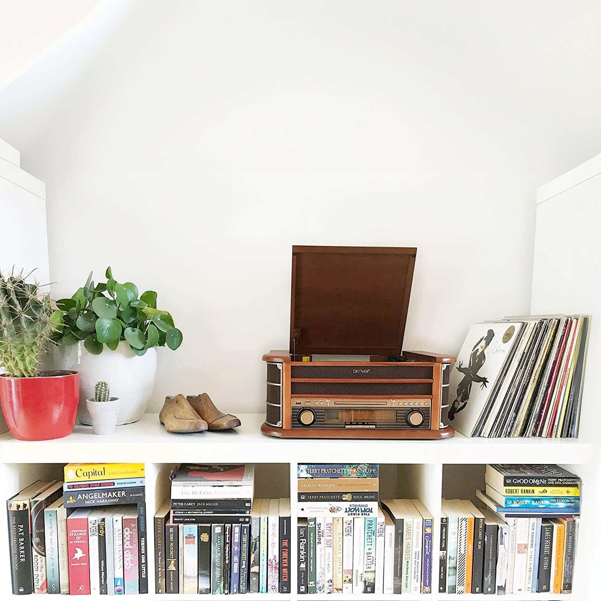 Een plank met boeken, planten en een vintage radio. De plank en de muur zijn wit. De radio is bruin.