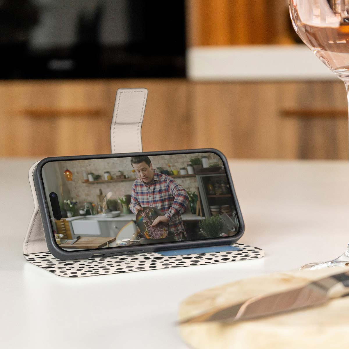 Een telefoonhoesje op een tafel met een video van een man die in een keuken kookt. Een glas wijn.