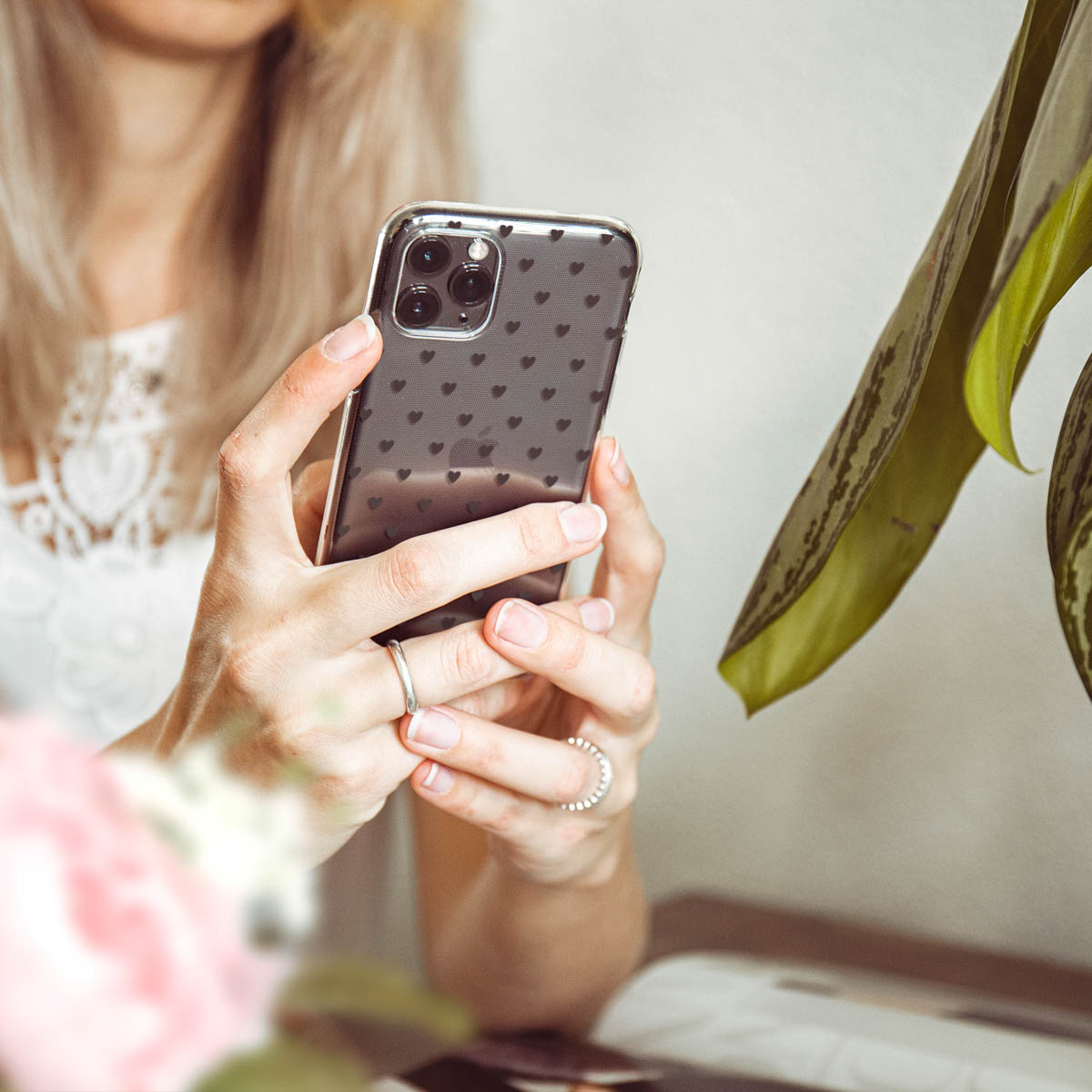 Een vrouw die een smartphone vasthoudt met een hoesje met hartjespatroon. Een plant en een ring zijn te zien.