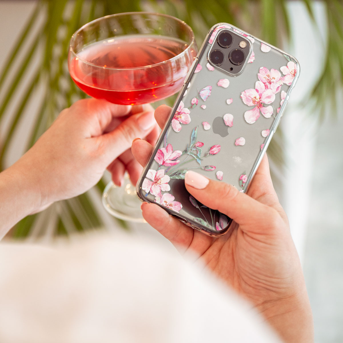 Een persoon die een telefoon met een bloemenhoes en een glas roze drank vasthoudt, met een groene plant op de achtergrond.