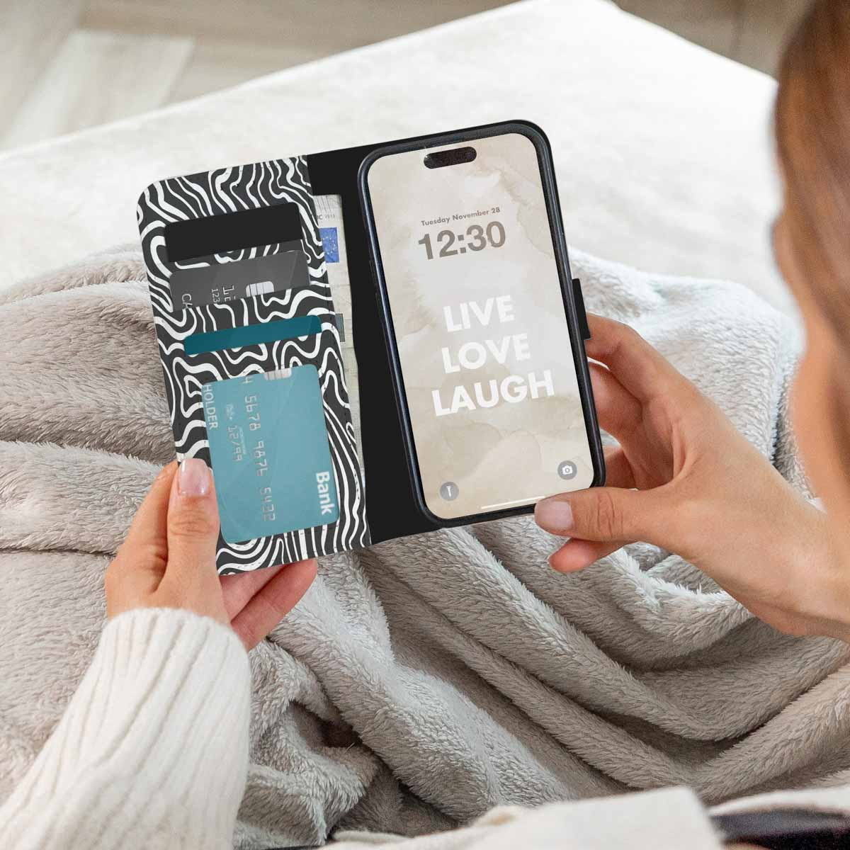 Een persoon houdt een telefoon vast met "Live, Love, Laugh" op het scherm en een portemonnee met kaarten. De persoon zit op een grijze deken.