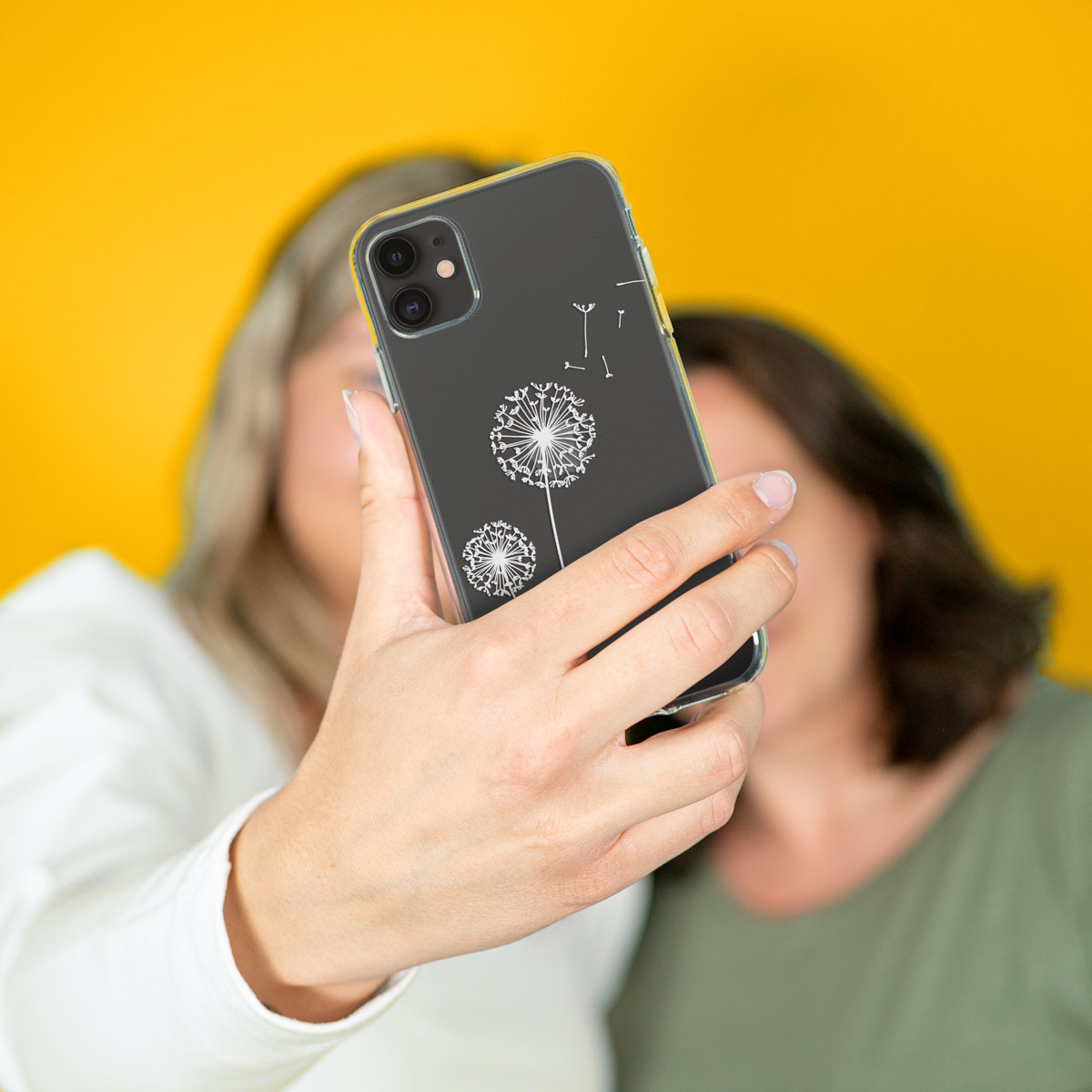 Twee vrouwen die een selfie maken met een telefoon in een doorzichtige hoes met een paardenbloemontwerp. De achtergrond is geel.