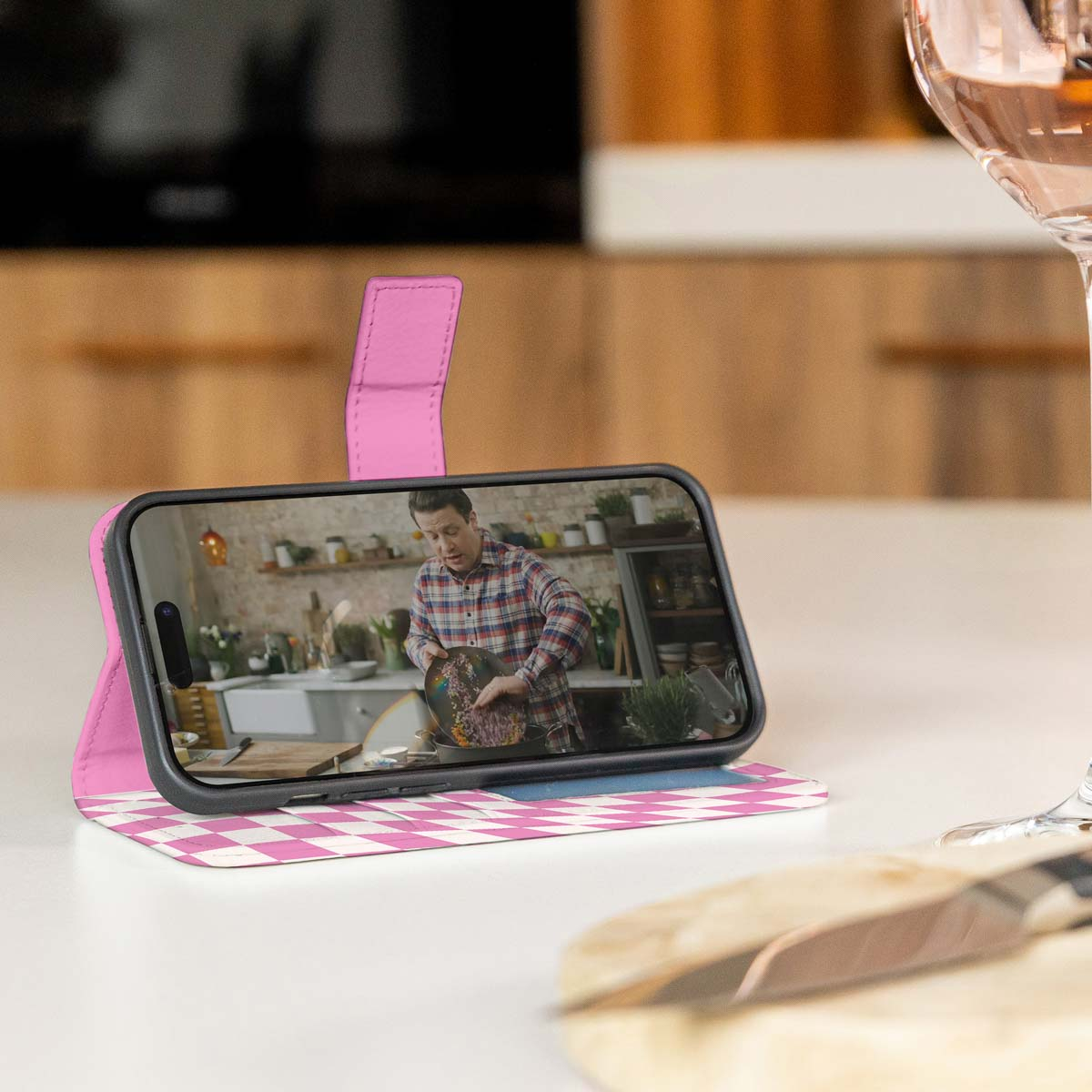 Telefoon in roze geruite hoes toont kok die kookt. Wijnglas aanwezig.