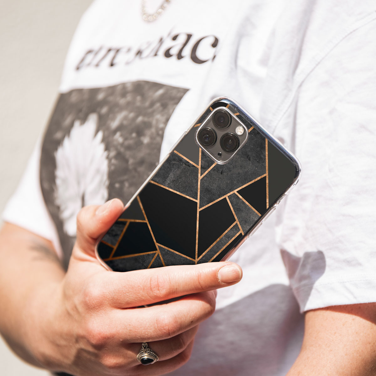 Personne tenant un téléphone avec un étui au design géométrique noir et or. La personne porte un t-shirt blanc.