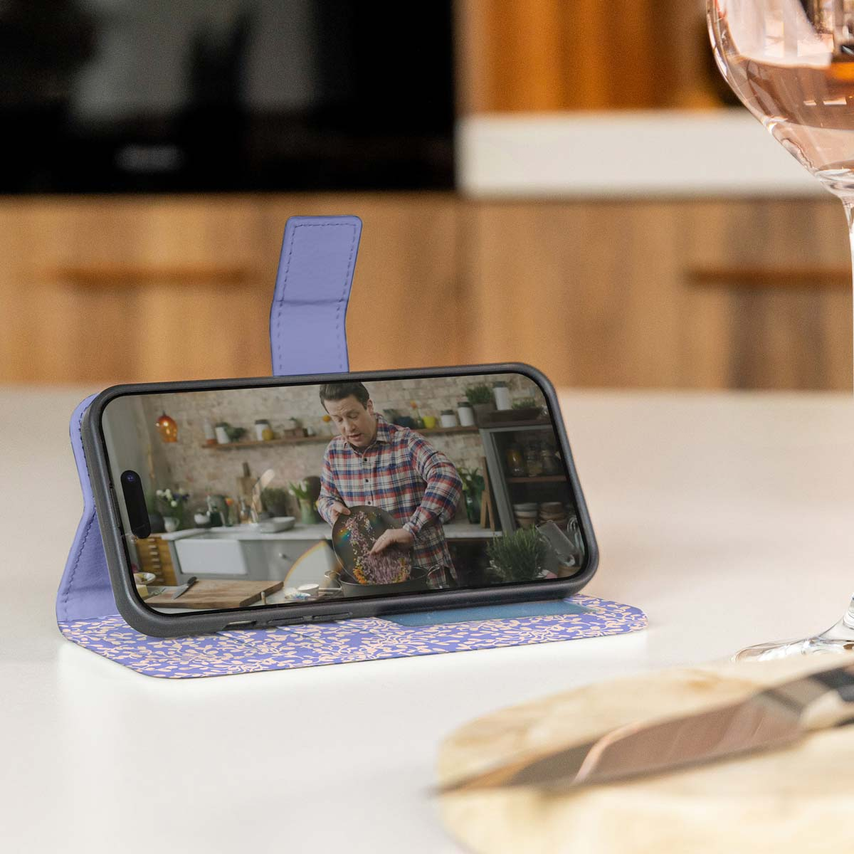 Een telefoon in een paarse hoes ligt op een tafel. Er staat een man op het scherm.