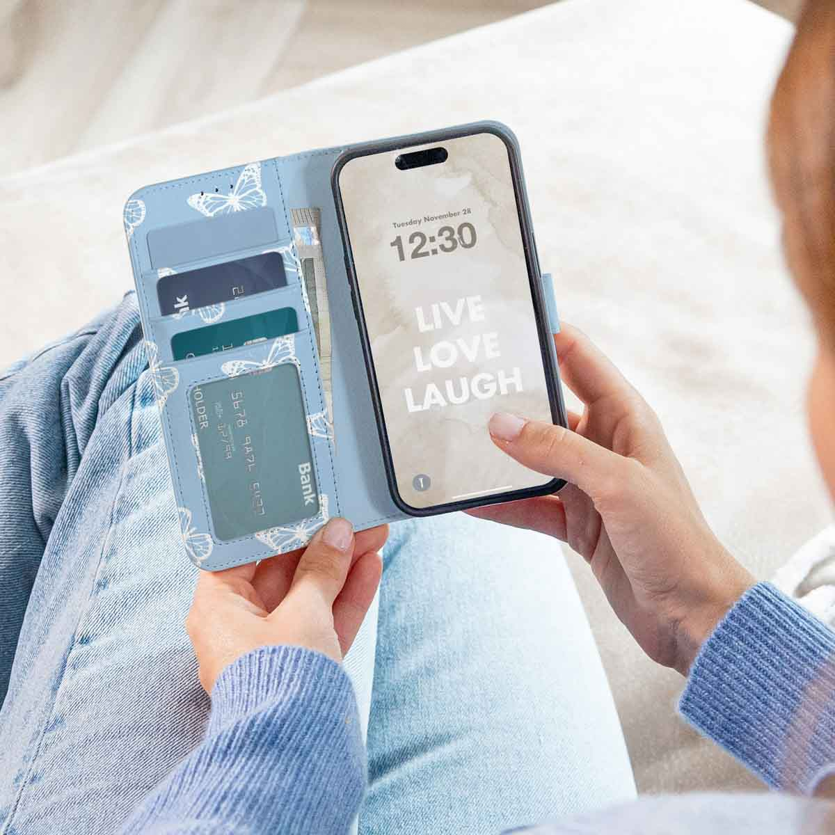 Een vrouw houdt een blauwe telefoonhoes met vlinders open. De telefoon toont 'Live, Love, Laugh'. Hoes heeft kaartsleuven.