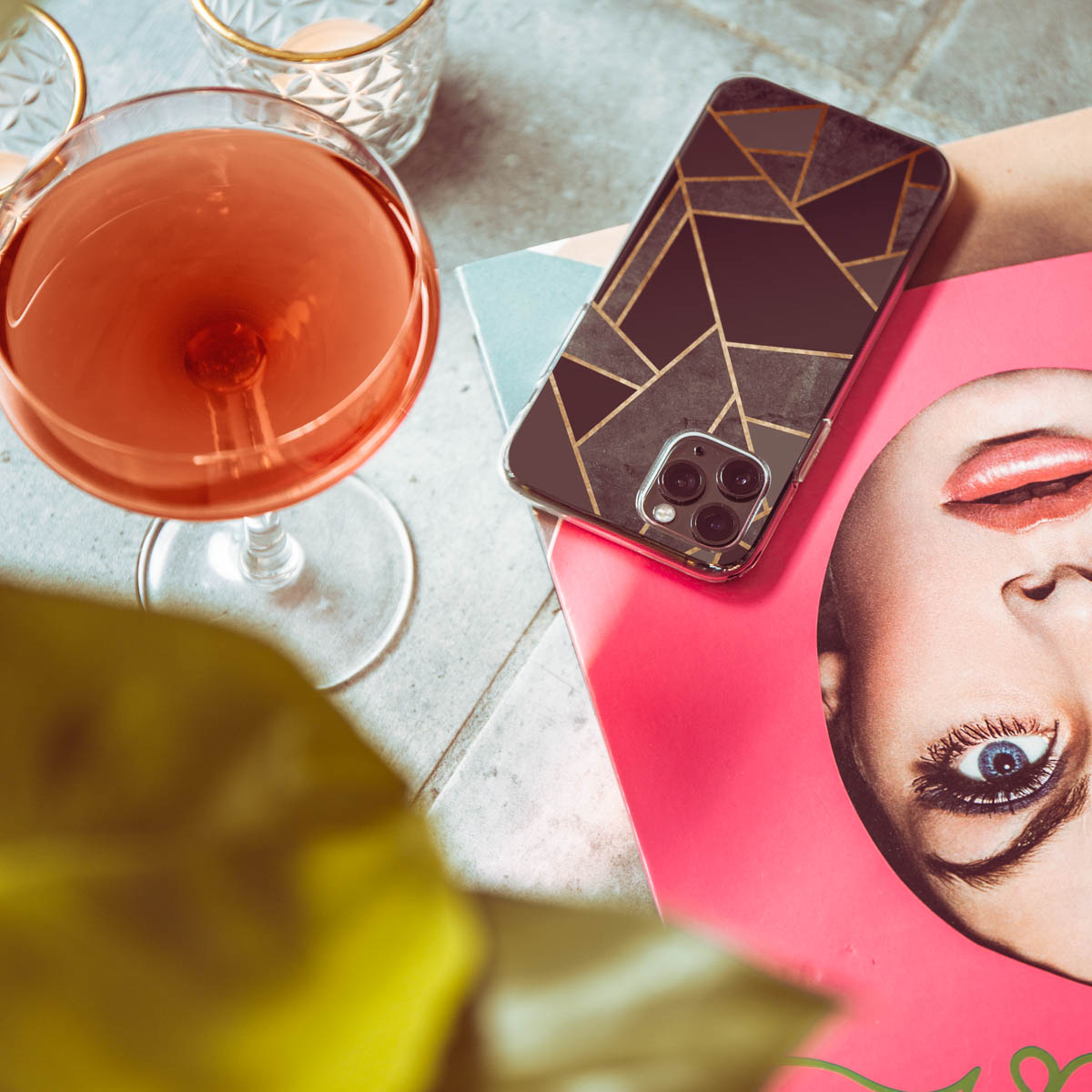Un téléphone avec un étui au design géométrique noir et or à côté d'un magazine rose, un verre de boisson rose et d'autres verres.