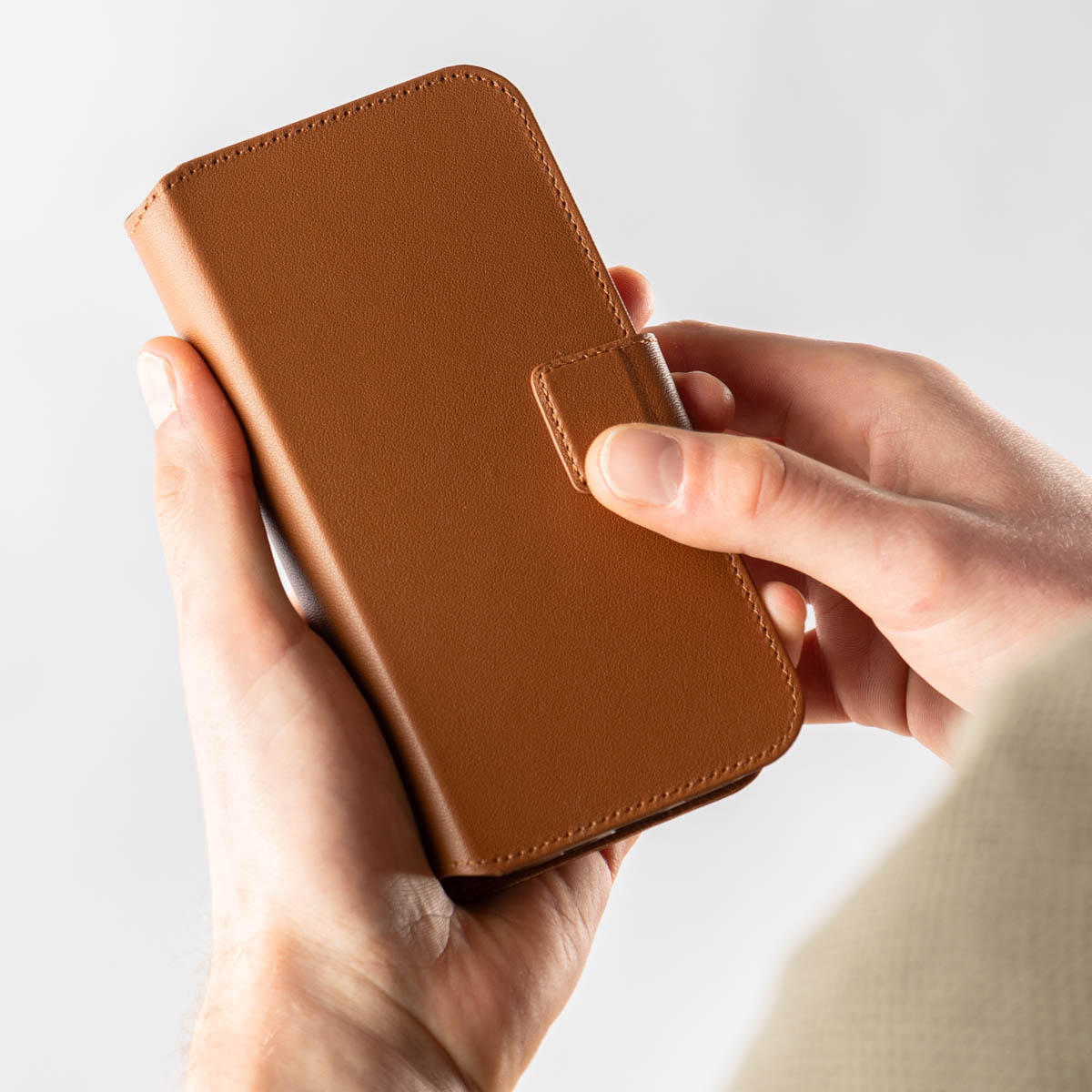 De handen van een persoon die een bruine leren telefoonhoes met een magnetische sluiting vasthoudt.