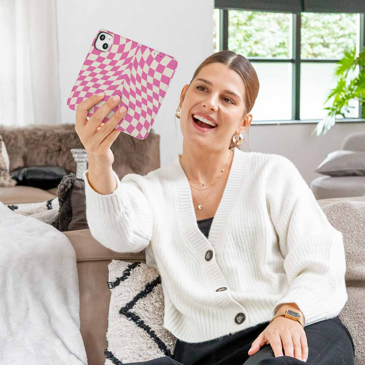 Vrouw die een telefoon vasthoudt met een roze en wit geblokte hoes. Ze lacht en draagt een witte trui.