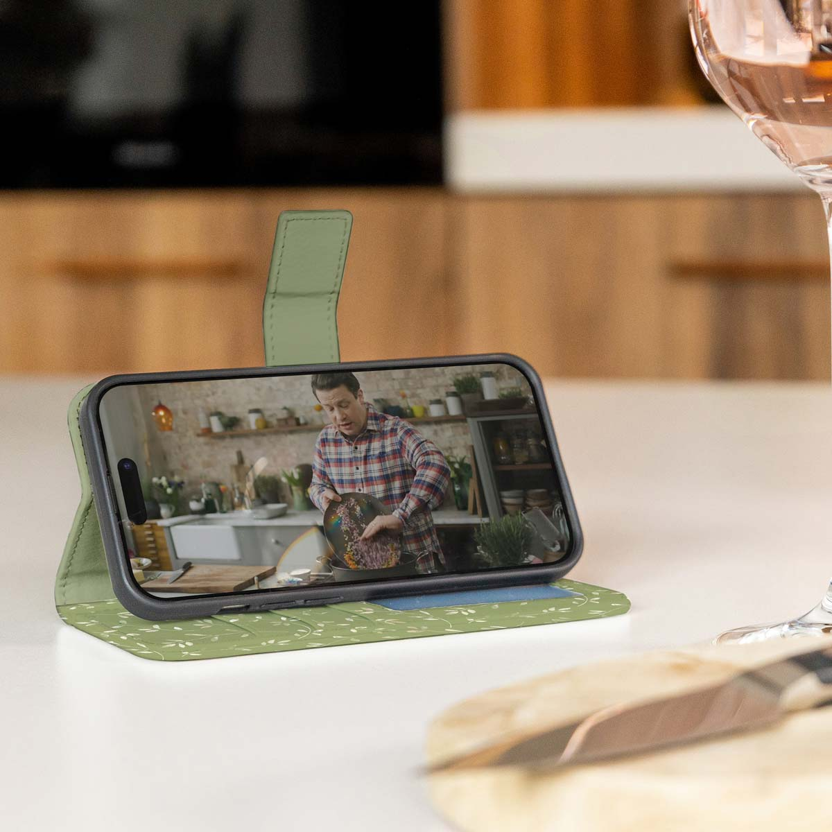 Telefoonhoesje met een telefoon die een kookvideo afspeelt, op een wit oppervlak, glas in de buurt.