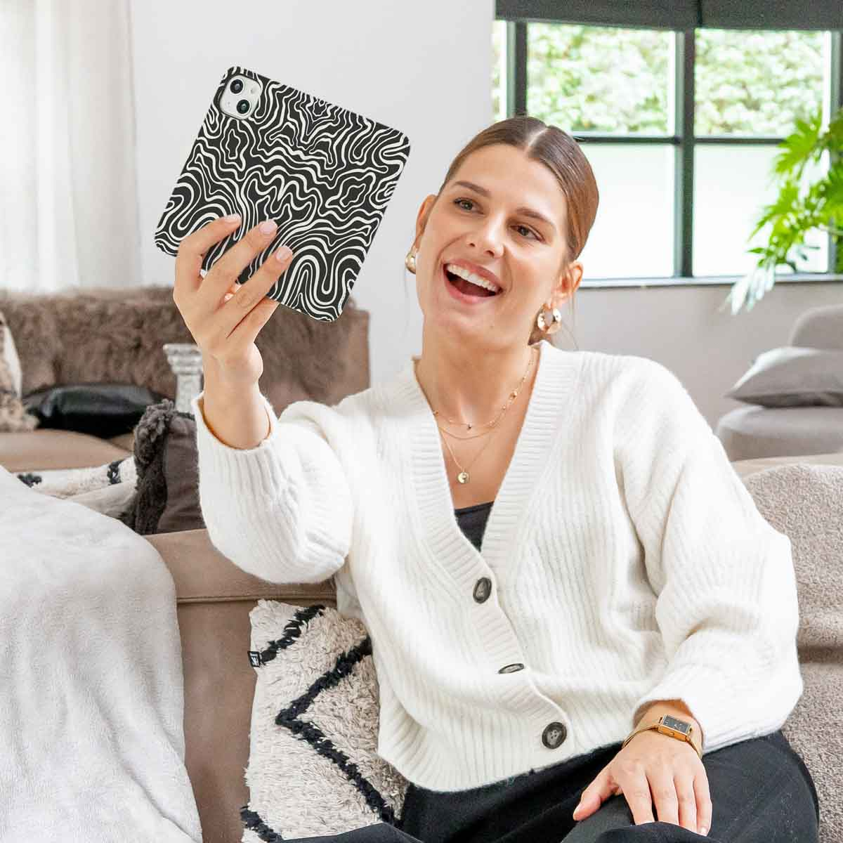 Een vrouw lacht en houdt een telefoonhoesje vast met een zwart-wit golfpatroon. Ze draagt een witte trui en zit op een bank.