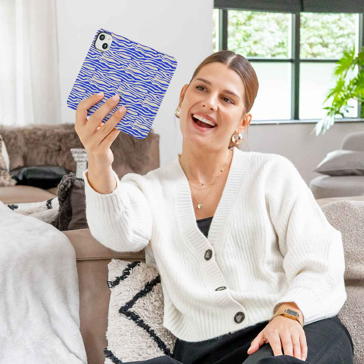 Vrouw houdt telefoon met blauw-witte zebra-gestreepte hoes vast, lachend. Achtergrond: bank, bloemen.