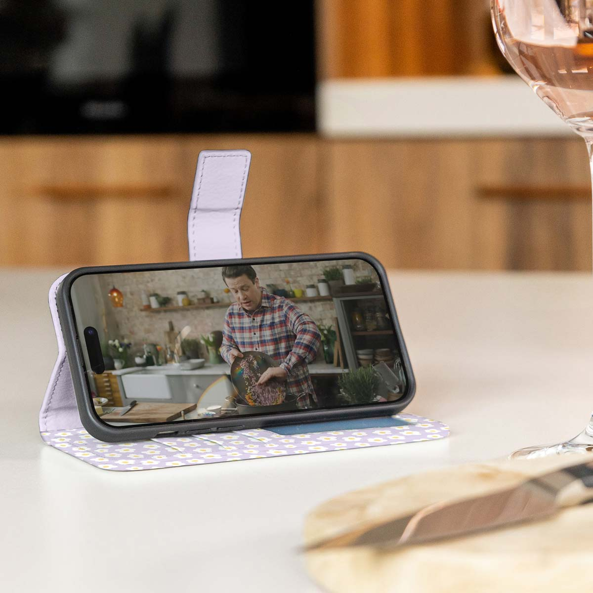 Telefoon in een paarse hoes, met een man die aan het koken is. Wijnglas rechts. Witte tafel.